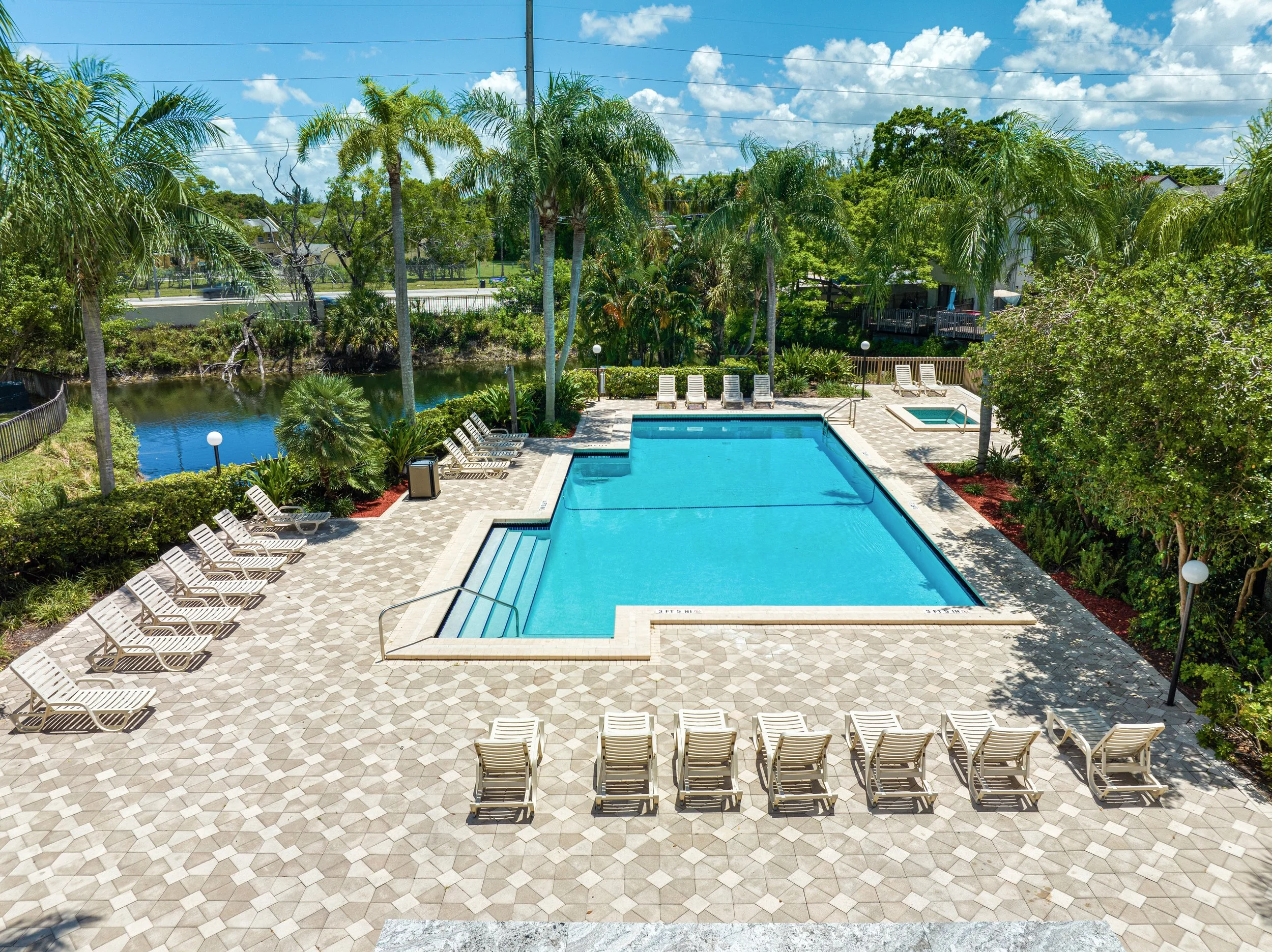A large swimming pool with a smaller pool attached, surrounded by lounge chairs and lush tropical trees and plants, in a sunny outdoor setting with blue sky and white clouds.