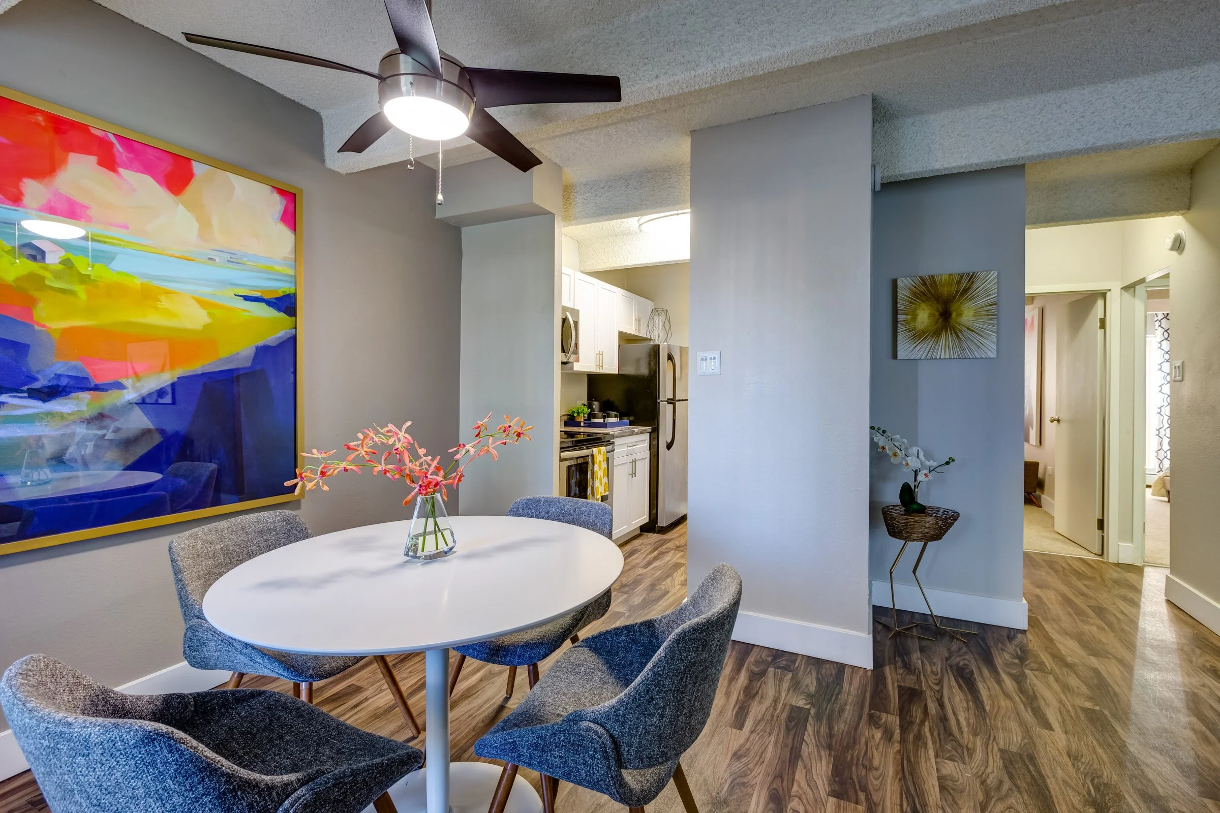 Interior view of a dining area with a white oval table and five gray upholstered chairs. A colorful abstract painting hangs on the wall, and a ceiling fan with a light is above. The background shows a kitchen with white cabinets and stainless steel a