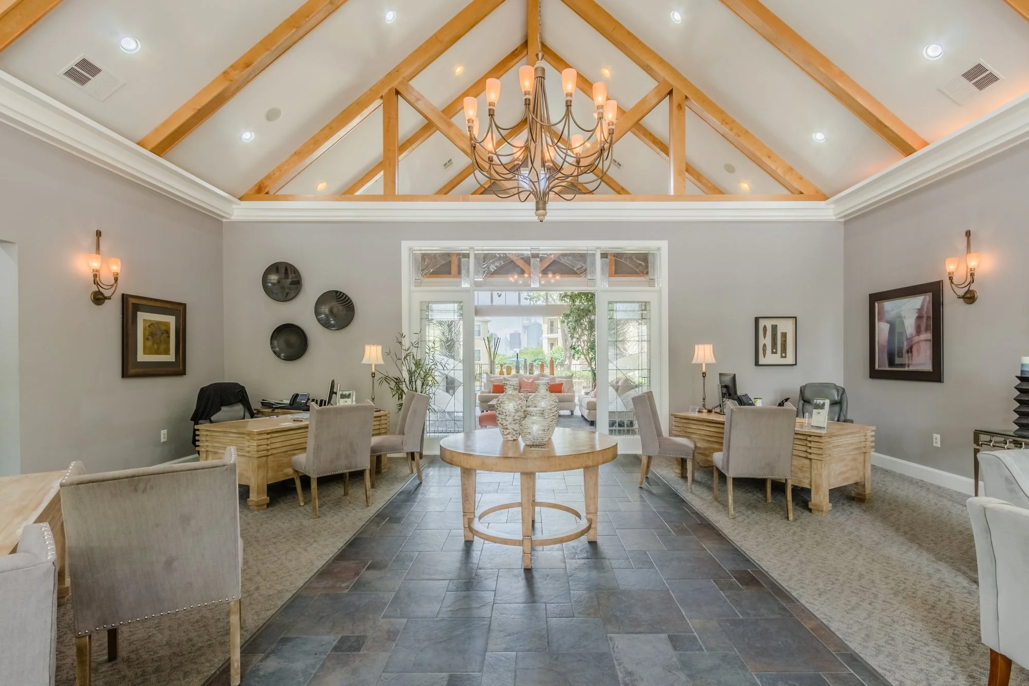 The image shows a spacious, well-lit lounge or reception area with a high vaulted ceiling featuring wooden beams and a large chandelier. There's a central round wooden table with decorative vases, surrounded by beige upholstered chairs. The back wall