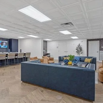 Modern lounge area with blue sofas, brown armchairs, coffee table with yellow and blue vases, and a potted plant. In the background, there are white walls, a door, and a long table with chairs.