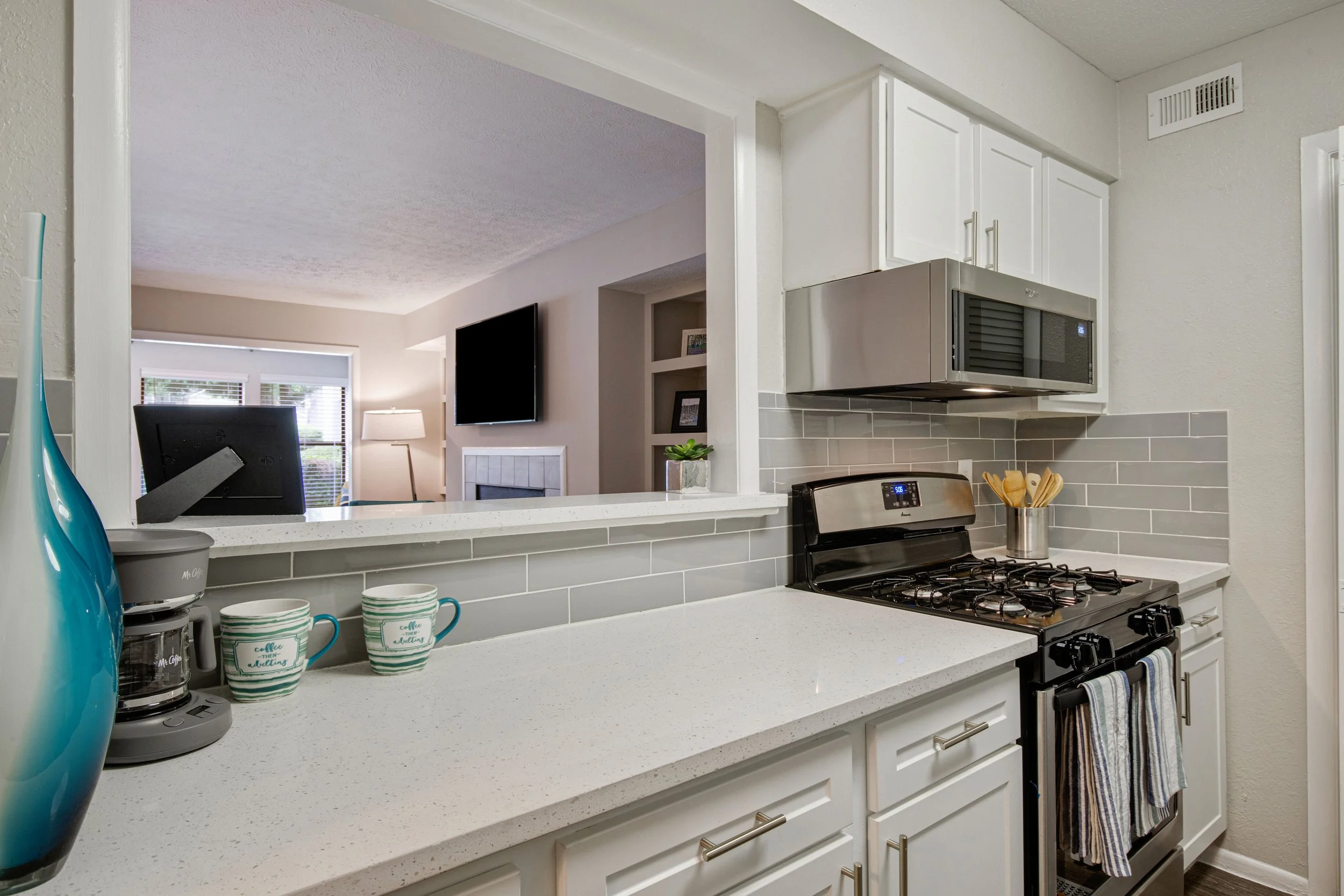 View of a kitchen with white cabinets, gray subway tile backsplash, a stainless steel microwave, and a gas stove. On the countertop, there is a coffee maker, two striped coffee cups, and a blue vase. A cut-out window opening looks into a living room 