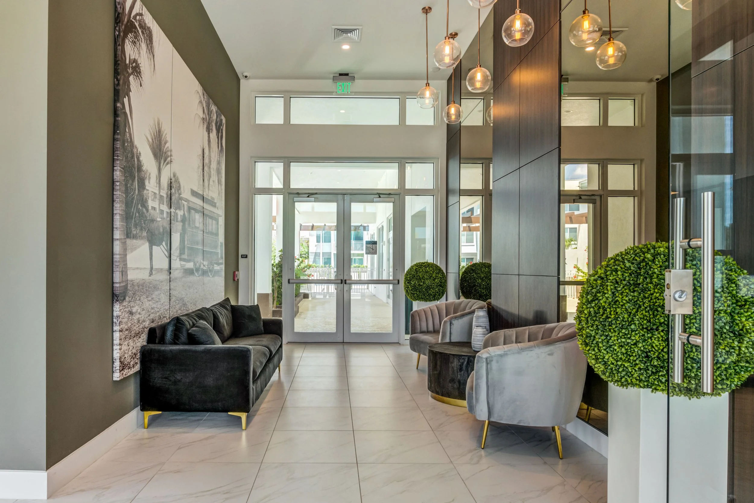 Modern hotel lobby with black and gray sofas, armchairs, large wall art of palm trees, glass pendant lights, and glass double doors leading outside.