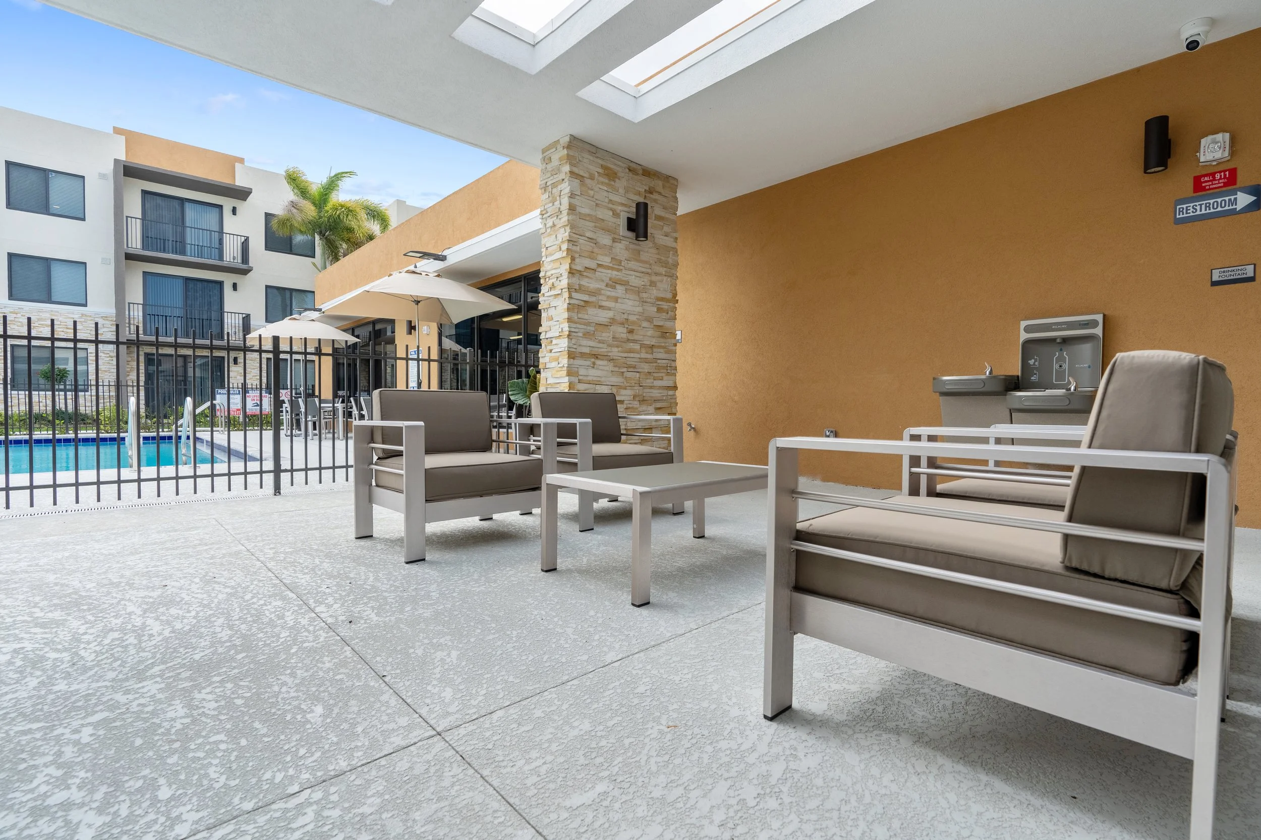Outdoor pool area with patio furniture, umbrellas, and a fountain, surrounded by apartment buildings.