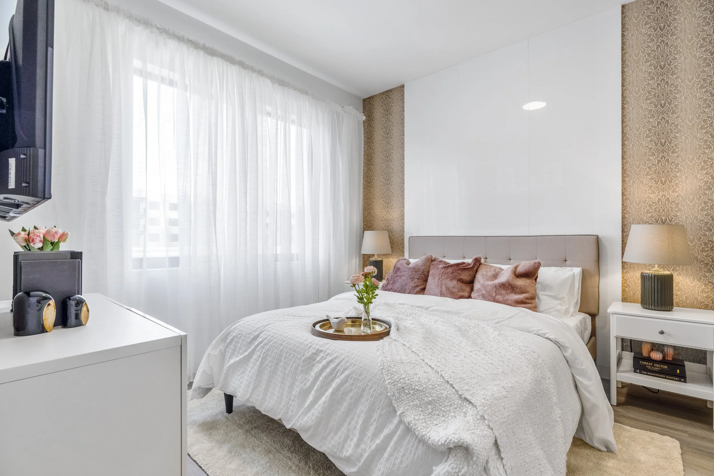 Bright bedroom with a large bed, pink pillows, white bedding, a tray with a flower on it, nightstands with lamps, and a window with sheer white curtains.