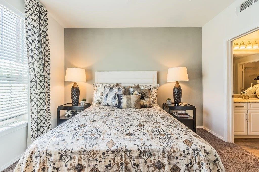 A neatly made bed with patterned bedding and multiple pillows in a bedroom with two matching lamps on black nightstands on either side, a window with patterned curtains, and an opening revealing a bathroom with white cabinetry.