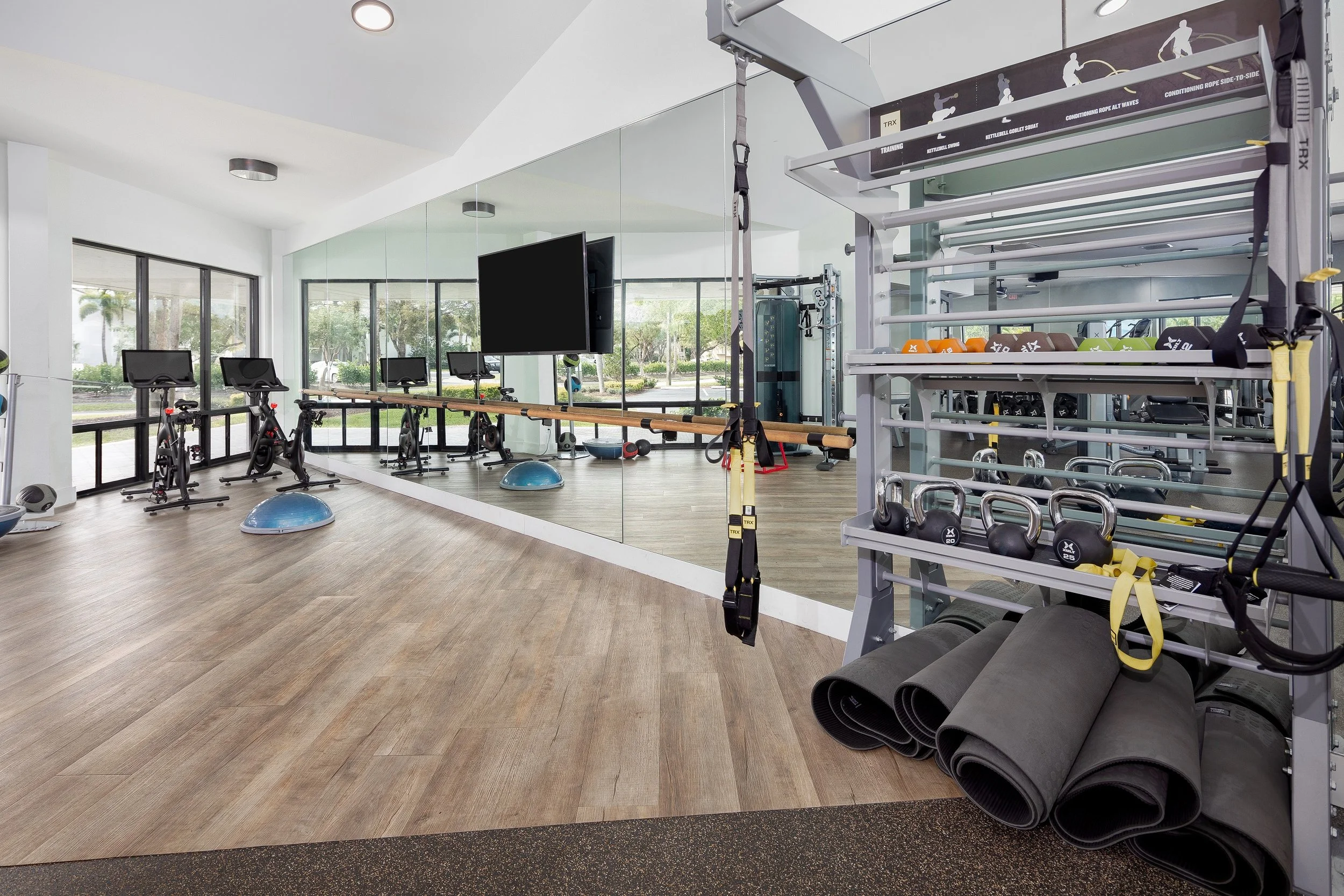 A modern gym with cardio equipment and a mirrored wall, featuring exercise mats, kettlebells, dumbbells, TRX suspension straps, and balance balls, with large windows showing an outdoor scene.