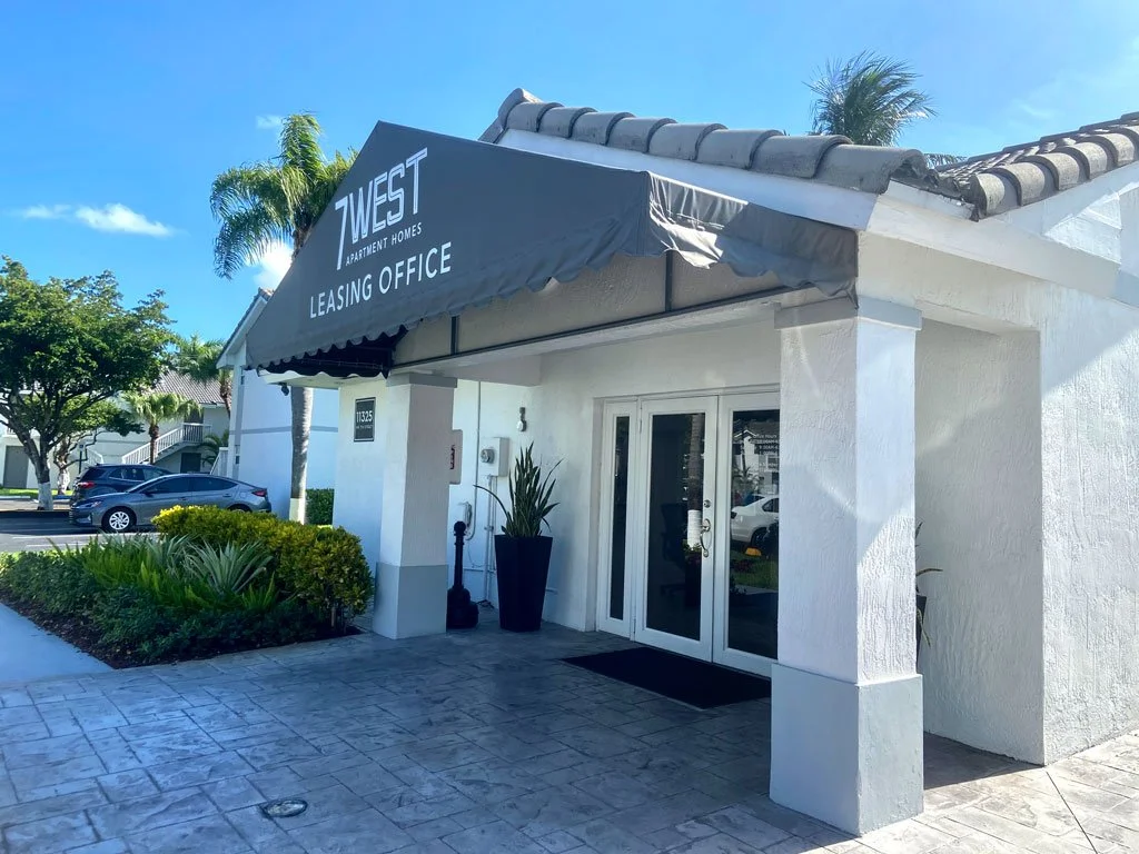 Exterior of a leasing office building with a black awning that reads "7 West Apartment Homes Leasing Office" above entrance, surrounded by palm trees and landscaping, with parked cars in background.