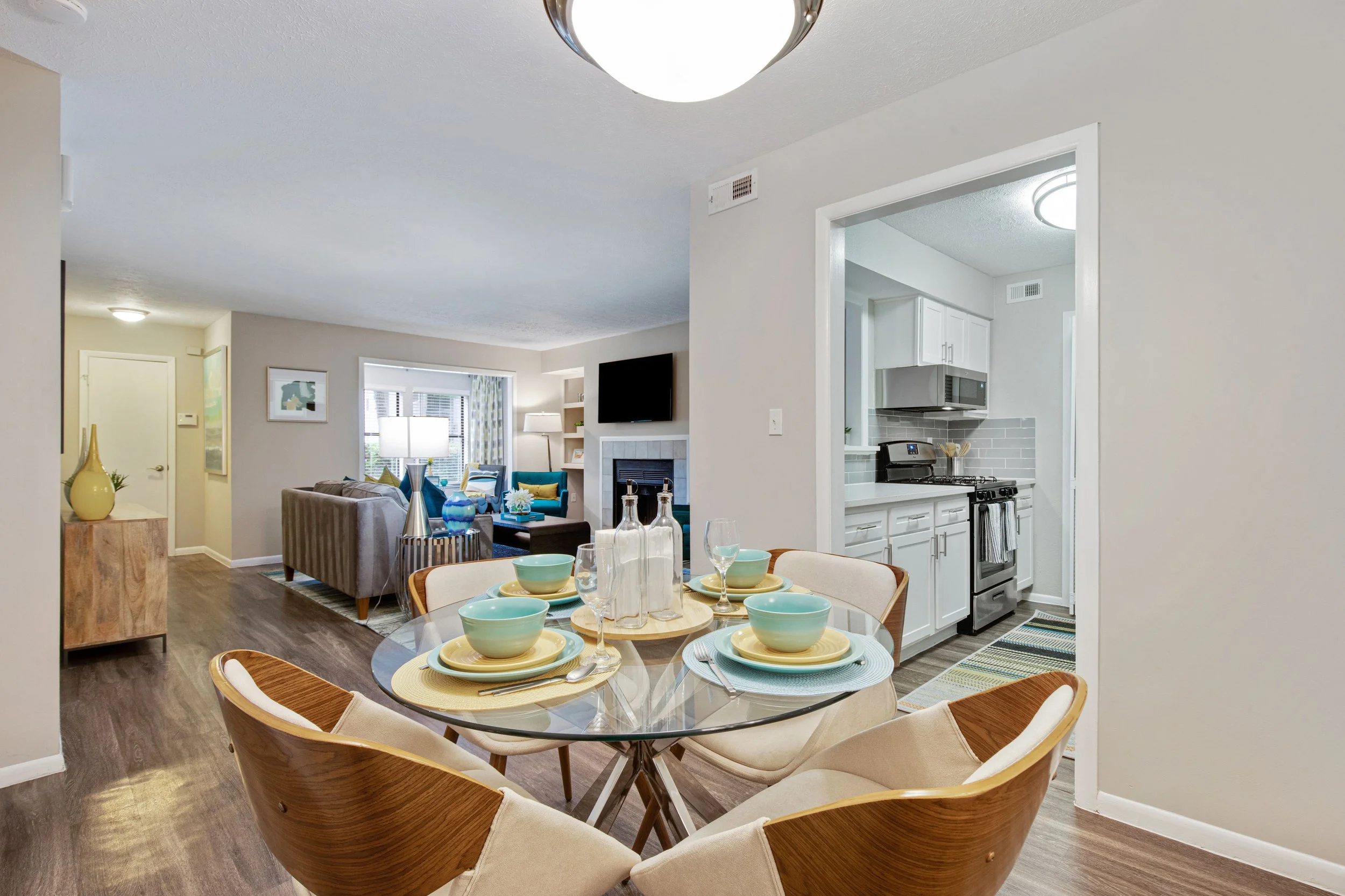 Open-concept living and dining area with view into kitchen. Dining table set with bowls, plates, glasses, and bottles, surrounded by mid-century modern chairs. Living room has beige sofa, armchairs, and a mounted TV above a fireplace.