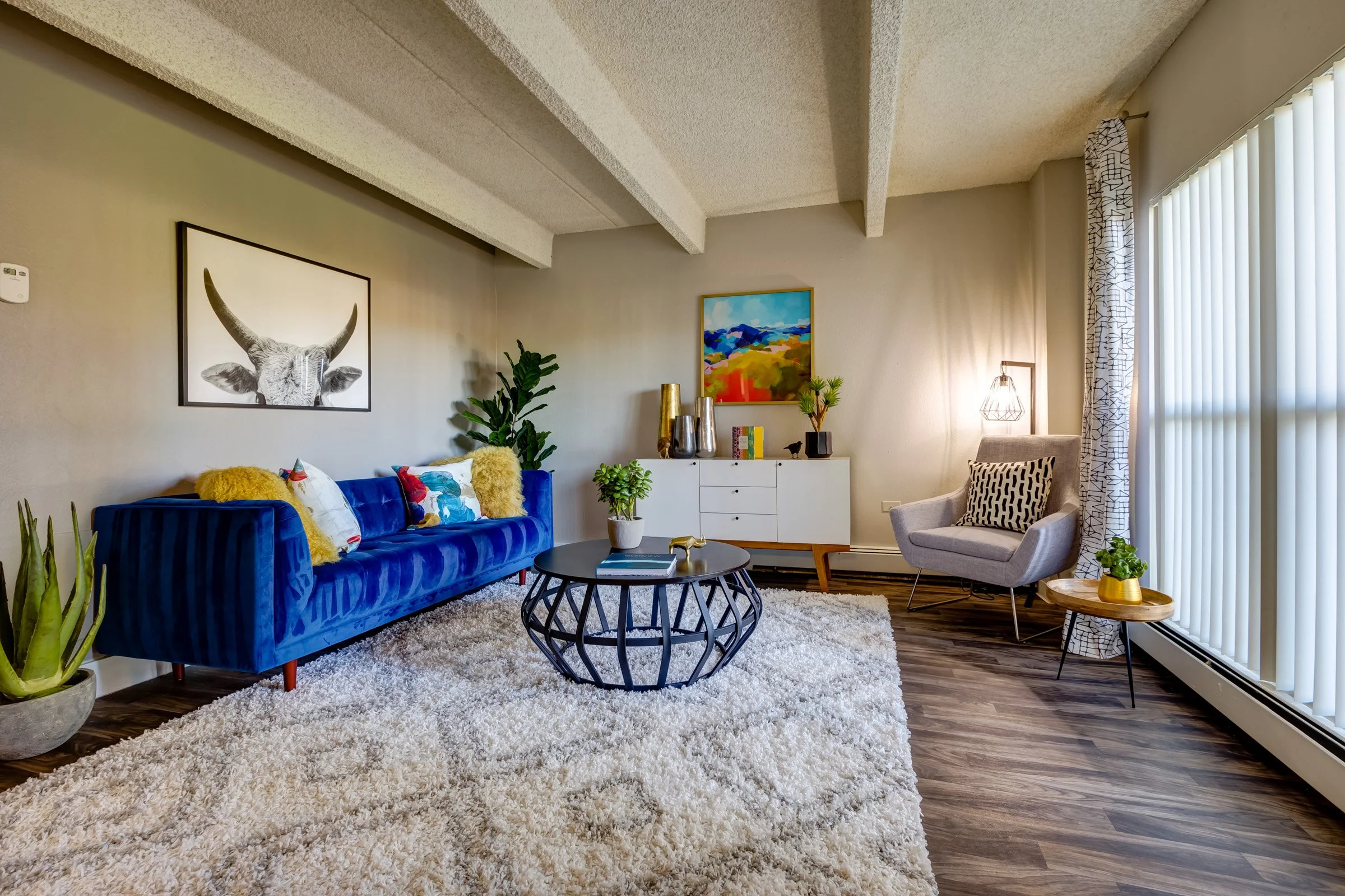 Living room with a blue velvet sofa, a beige armchair with a black and white pillow, a round black coffee table, a white sideboard with decorative vases and books, a potted plant, and modern artwork on the walls. Large window with vertical blinds and