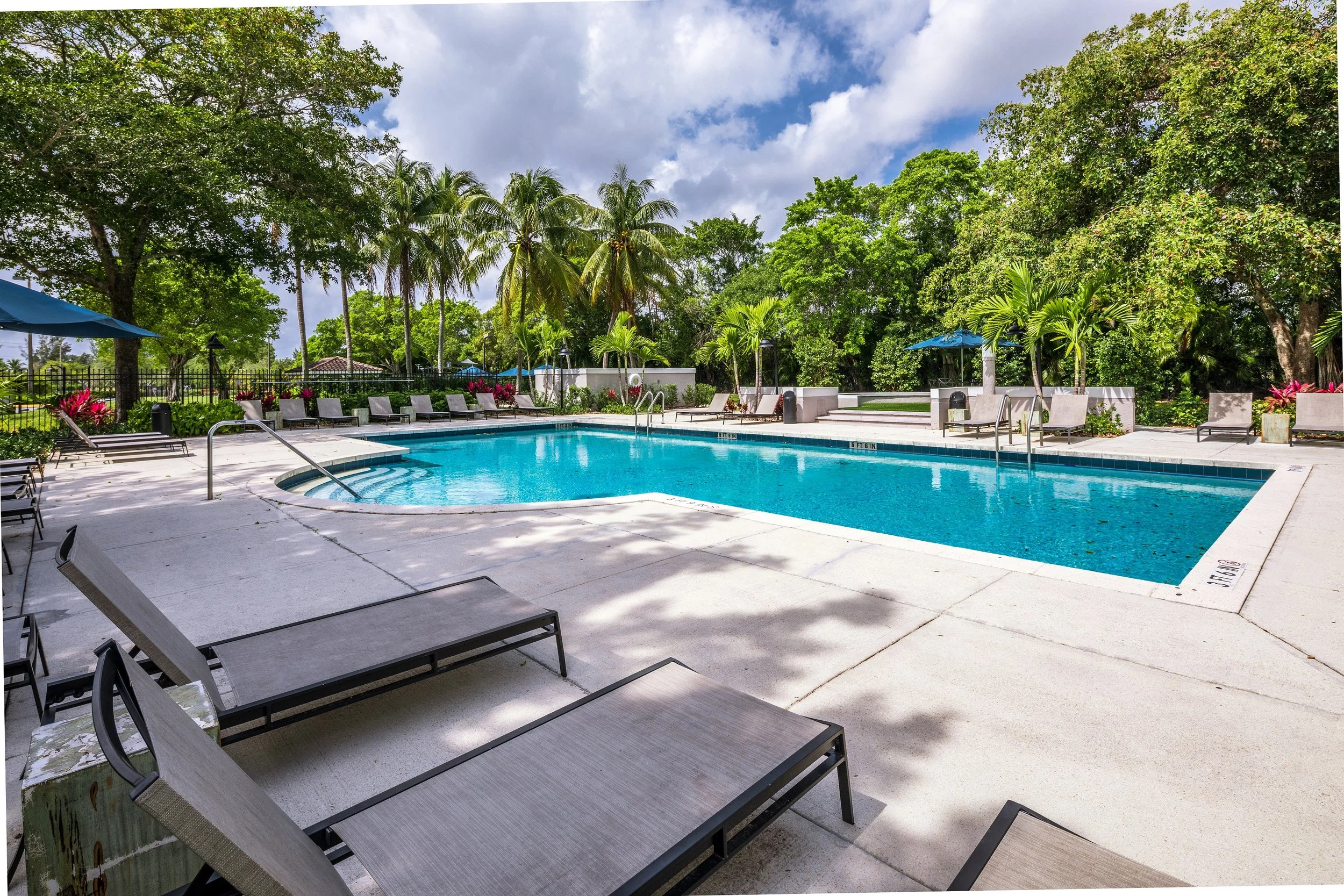 Outdoor swimming pool surrounded by lounge chairs, umbrellas, and lush green trees under a partly cloudy sky.