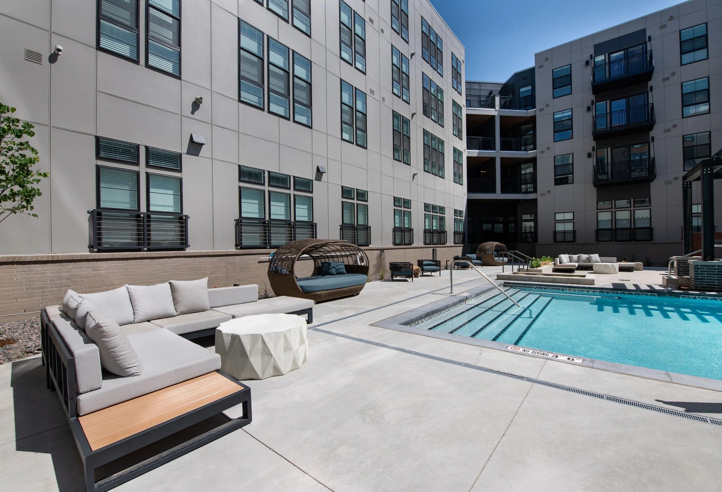 Apartment complex courtyard with swimming pool and outdoor seating area, including a brown wicker daybed, white couches, and various chairs, surrounded by modern multi-story buildings.