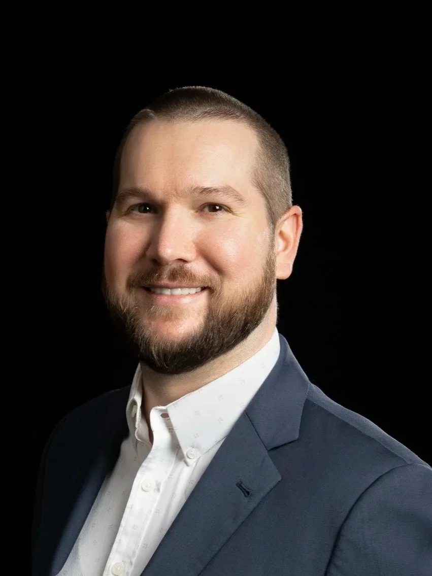 Professional headshot of a young man with short brown hair, a full beard, wearing a navy suit, light blue dress shirt, and a striped tie, smiling against a dark background.