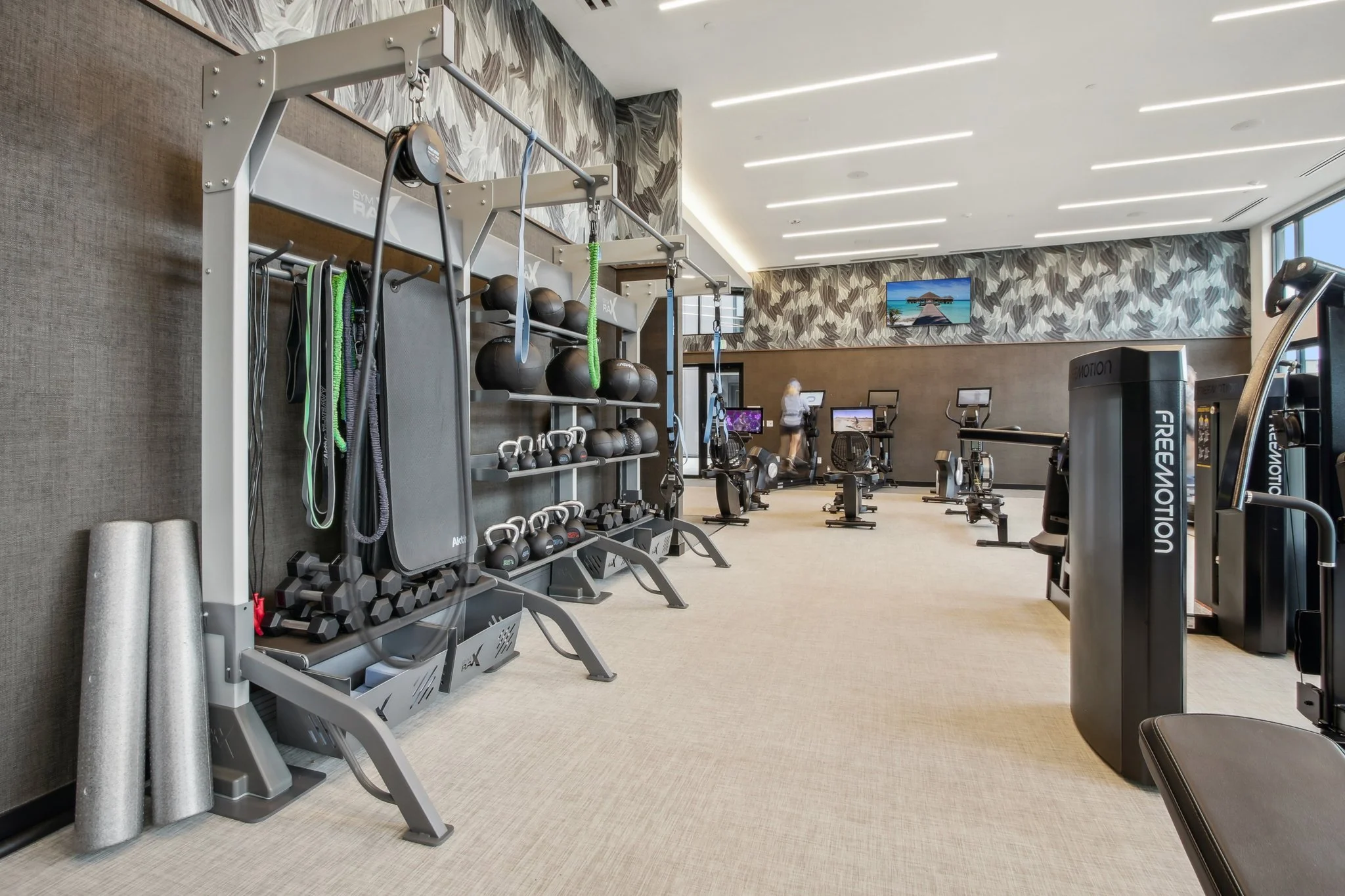 Empty modern gym with exercise equipment including kettlebells, medicine balls, and resistance bands, with a woman using a stationary bike in the background.