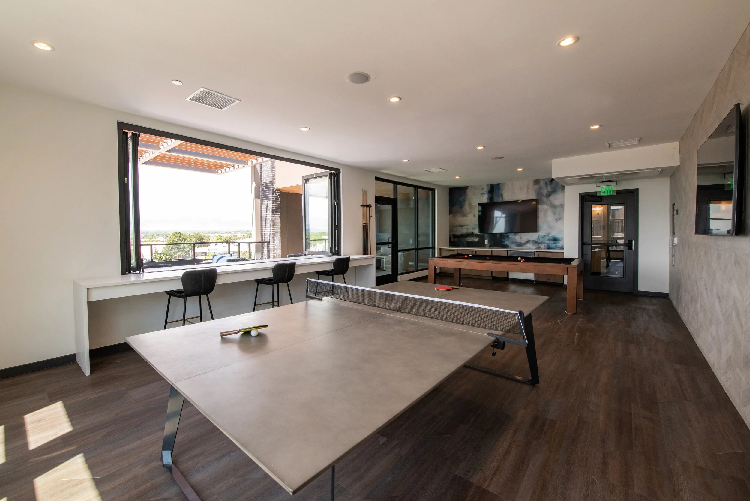 Indoor recreational room with ping pong table, foosball table, wall-mounted TV, window view, and bar seating.