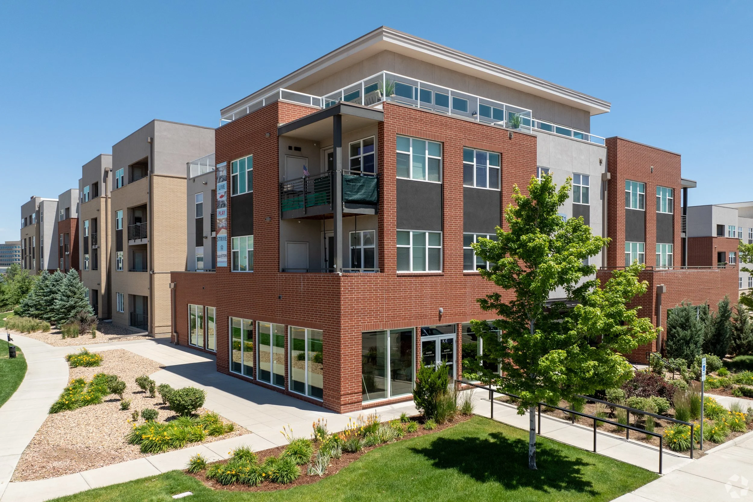 Modern multi-story apartment building with brick, concrete, and glass exterior, surrounded by landscaped walkways, trees, and greenery under a clear blue sky.