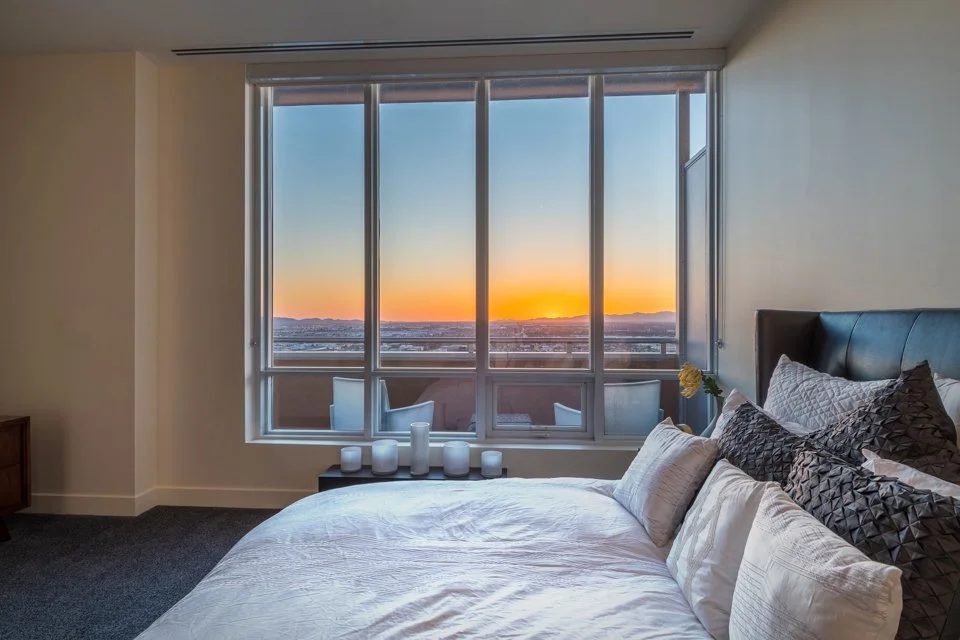 A bedroom with a large window showing a sunset over a city skyline. The bed has white and dark gray pillows, and white bedding. Candles are placed on a small table near the window, and a single flower is in a vase on the bedside table.