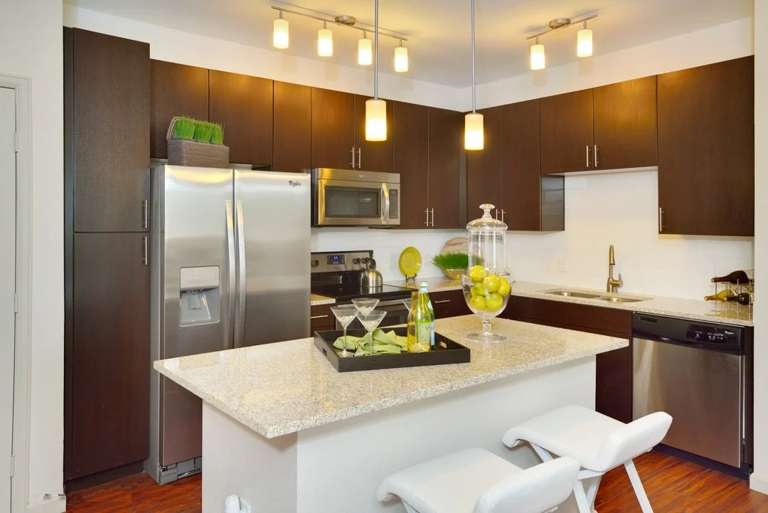 Modern kitchen with dark wood cabinets, stainless steel refrigerator, microwave, and dishwasher. A white island with a granite countertop has a tray with drinks and a glass jar filled with green apples. Pendant lights hang from the ceiling, and the k