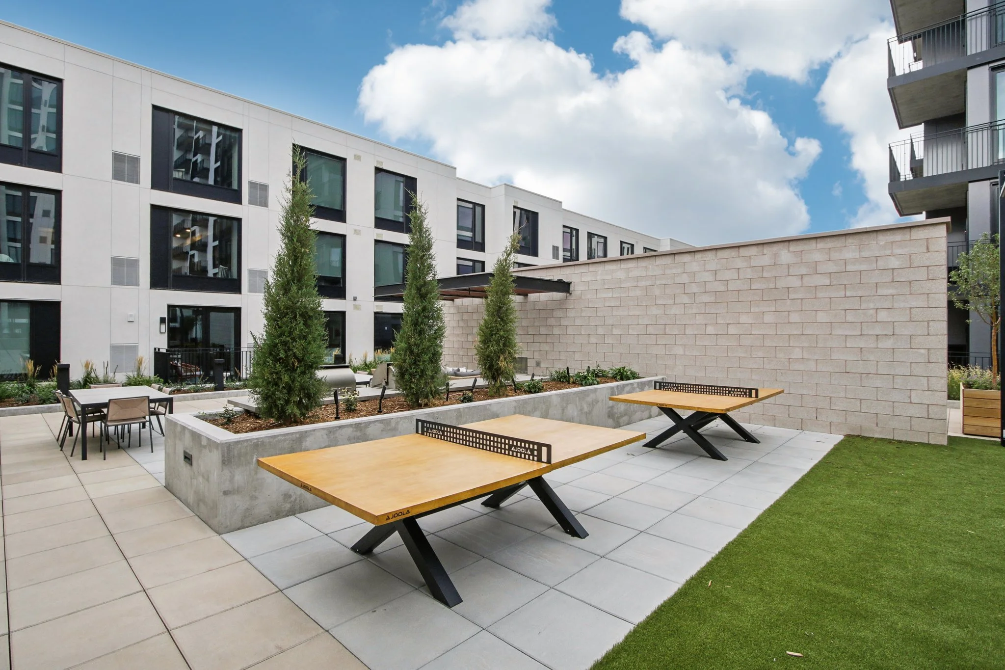 Outdoor patio with two ping pong tables, a small dining table with four chairs, tall trees in a raised planter, a brick wall, and an apartment building in the background under a partly cloudy sky.