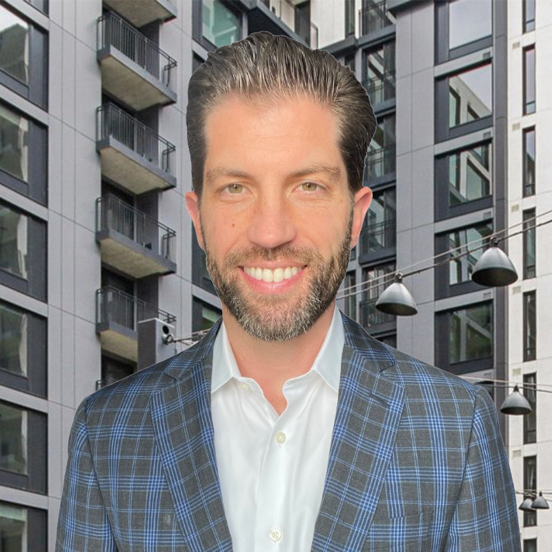 A man with a beard and slicked-back hair smiling, wearing a white shirt and a blue plaid blazer, standing in front of a modern apartment building.