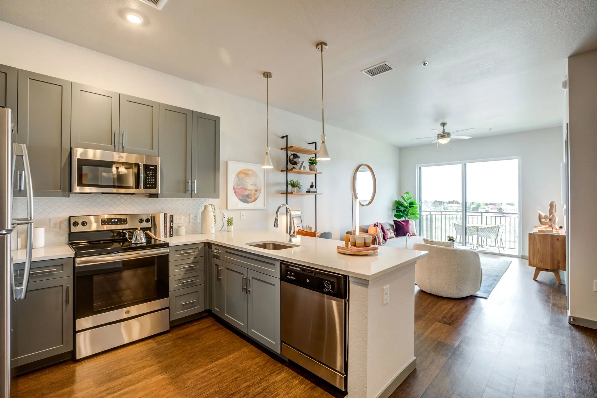 Open-plan kitchen and living room with gray cabinets, stainless steel appliances, wooden flooring, a balcony with outdoor seating, and contemporary decor with plants and artwork.