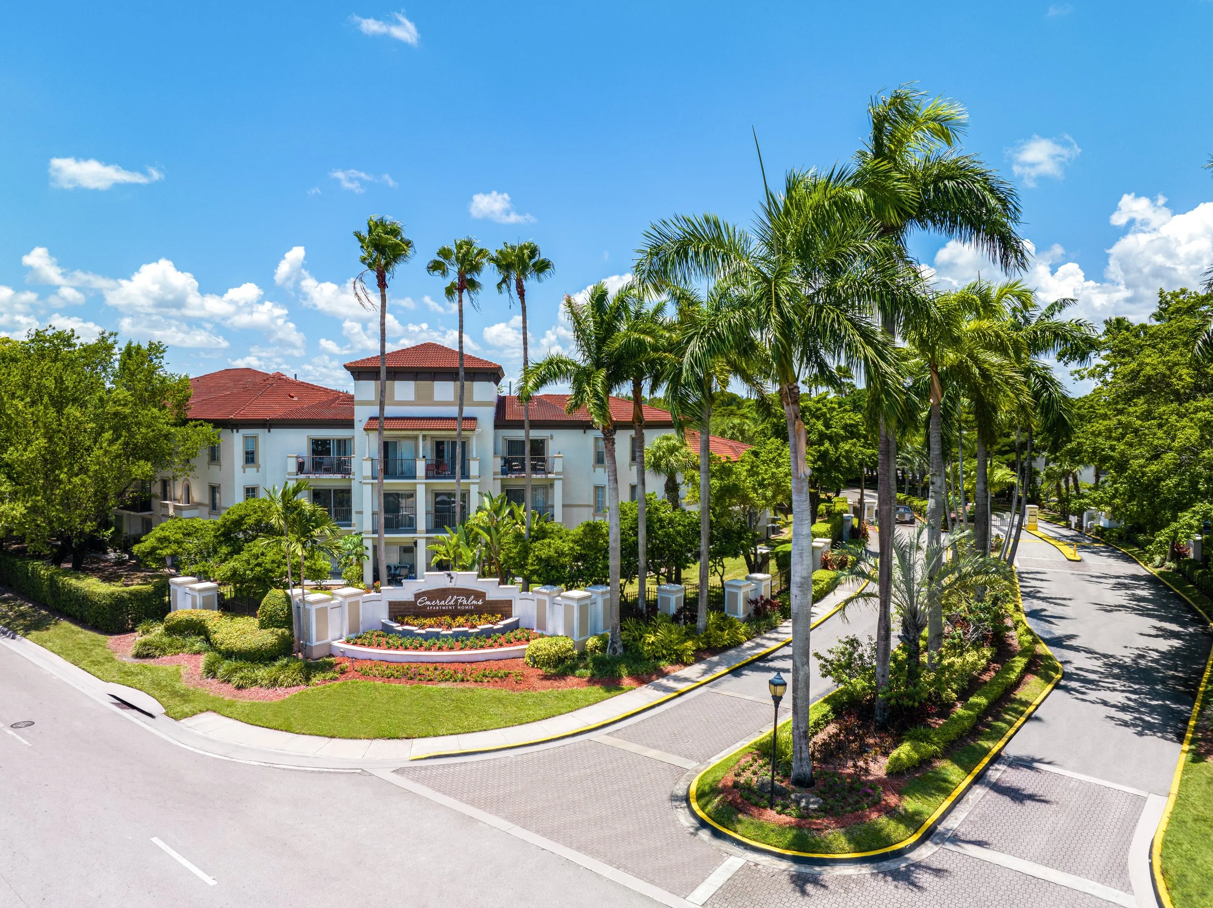 Gated apartment complex entrance with lush landscaping, tall palm trees, and a multi-story building with white walls and a red roof under a partly cloudy blue sky.
