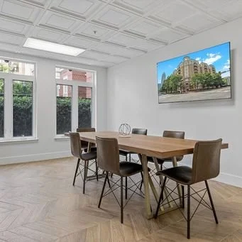 Empty conference room with a wooden table, six chairs, a large TV screen on the wall displaying a building, and windows showing trees outside.