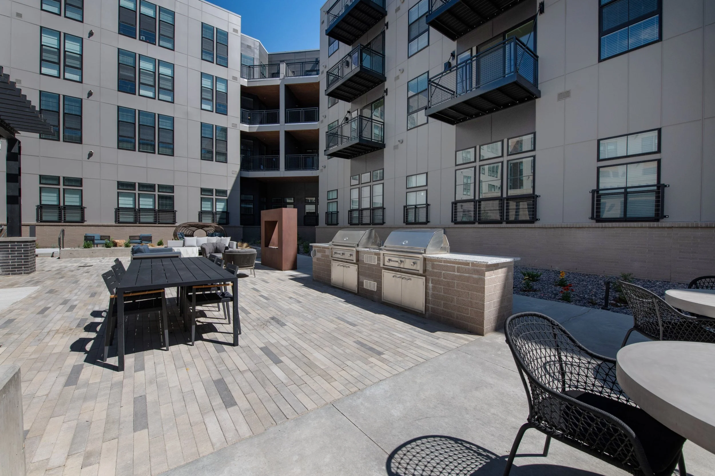 Outdoor apartment courtyard with lounge furniture, a dining table with chairs, built-in grills, and modern apartment building with multiple balconies