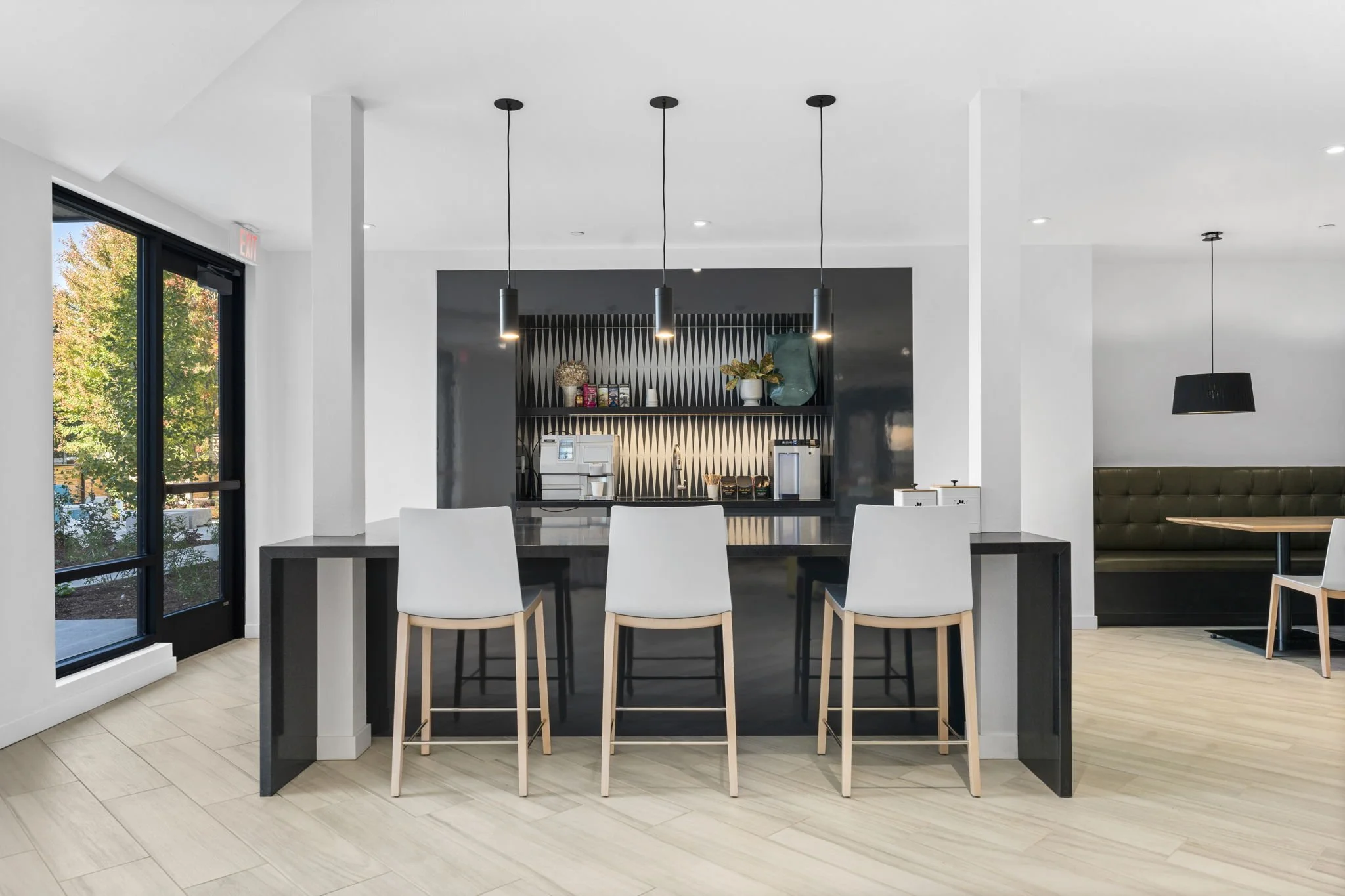 Modern kitchen with a breakfast bar, three white chairs, black pendant lights, a black and white patterned backsplash, and a coffee station.