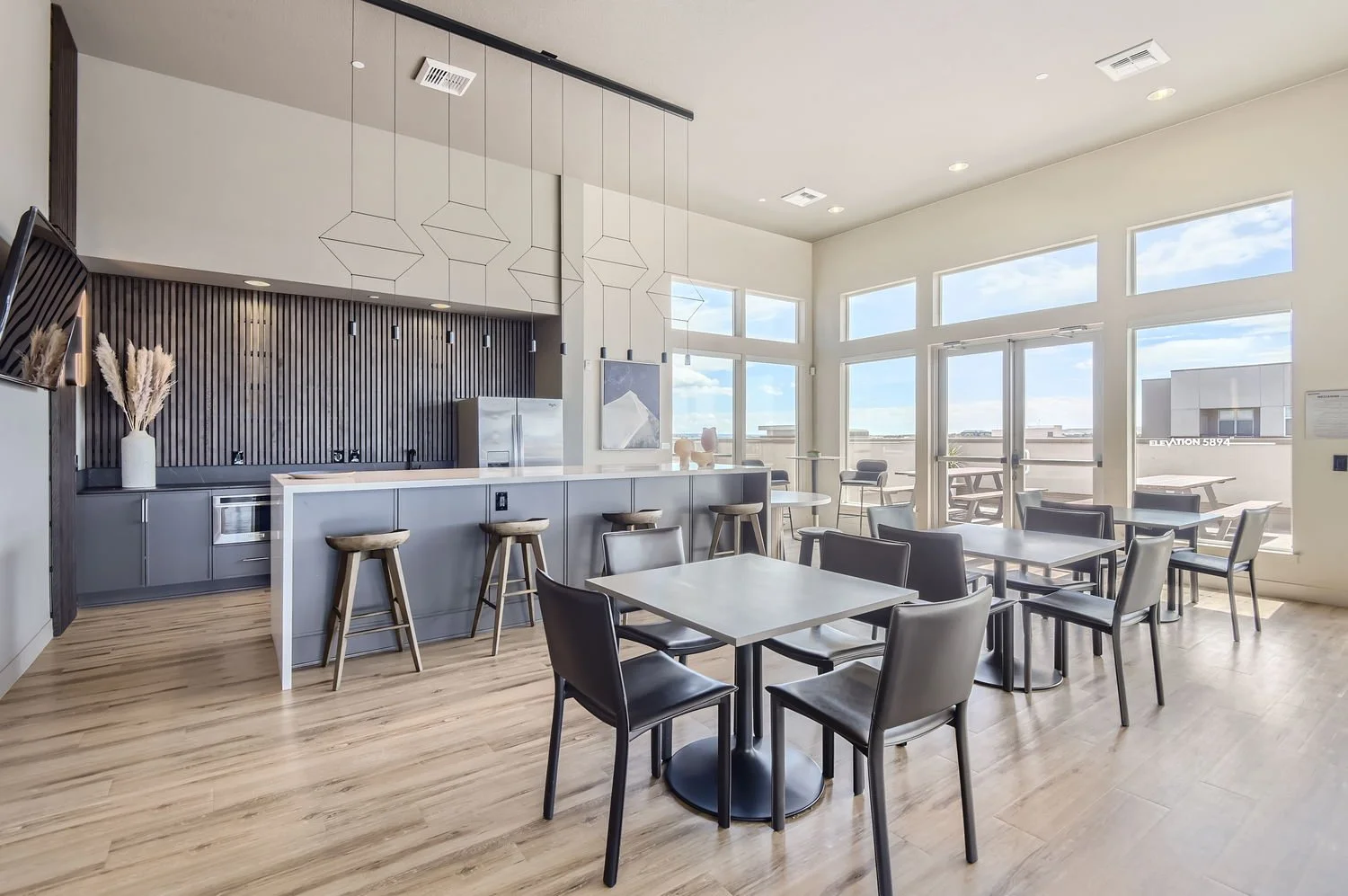 A modern, bright cafe or lounge area with a kitchenette, bar stools, tables, and chairs, featuring large windows and natural light.