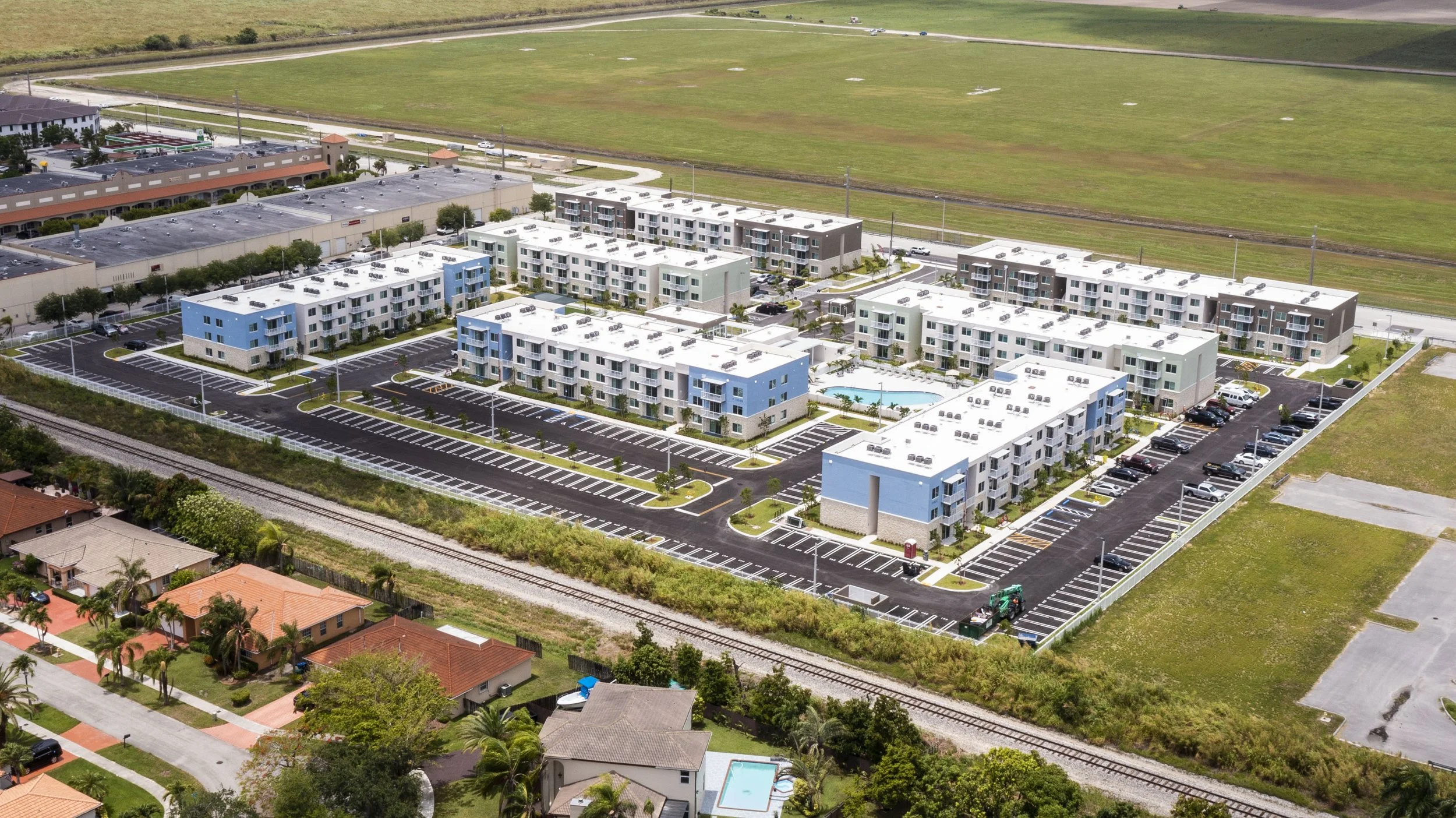Aerial view of a modern apartment complex with multiple buildings around a swimming pool and parking lot, adjacent to a railway track and neighboring houses.