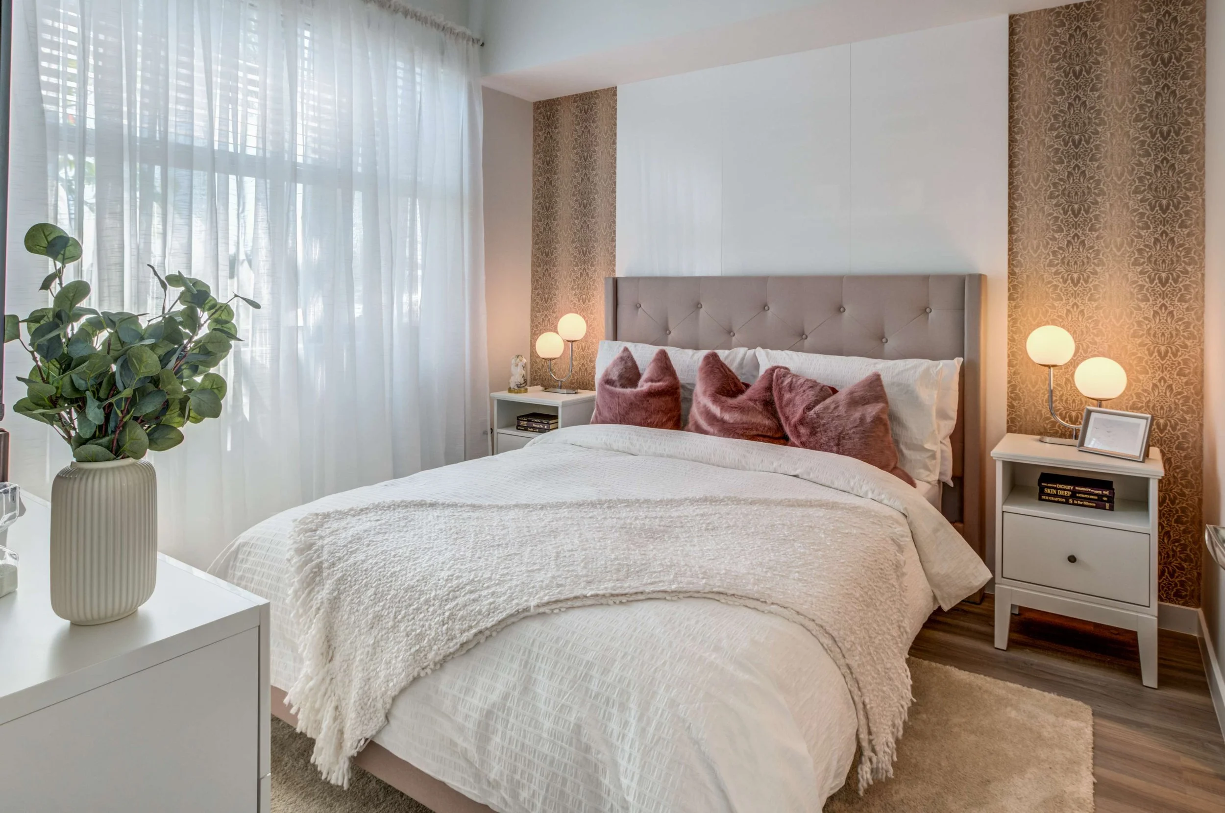 A modern bedroom with a large bed featuring a beige upholstered headboard and white bedding, decorated with pink velvet pillows. Flanking the bed are white nightstands with round lamps, and a potted plant is placed on a white dresser near a window wi