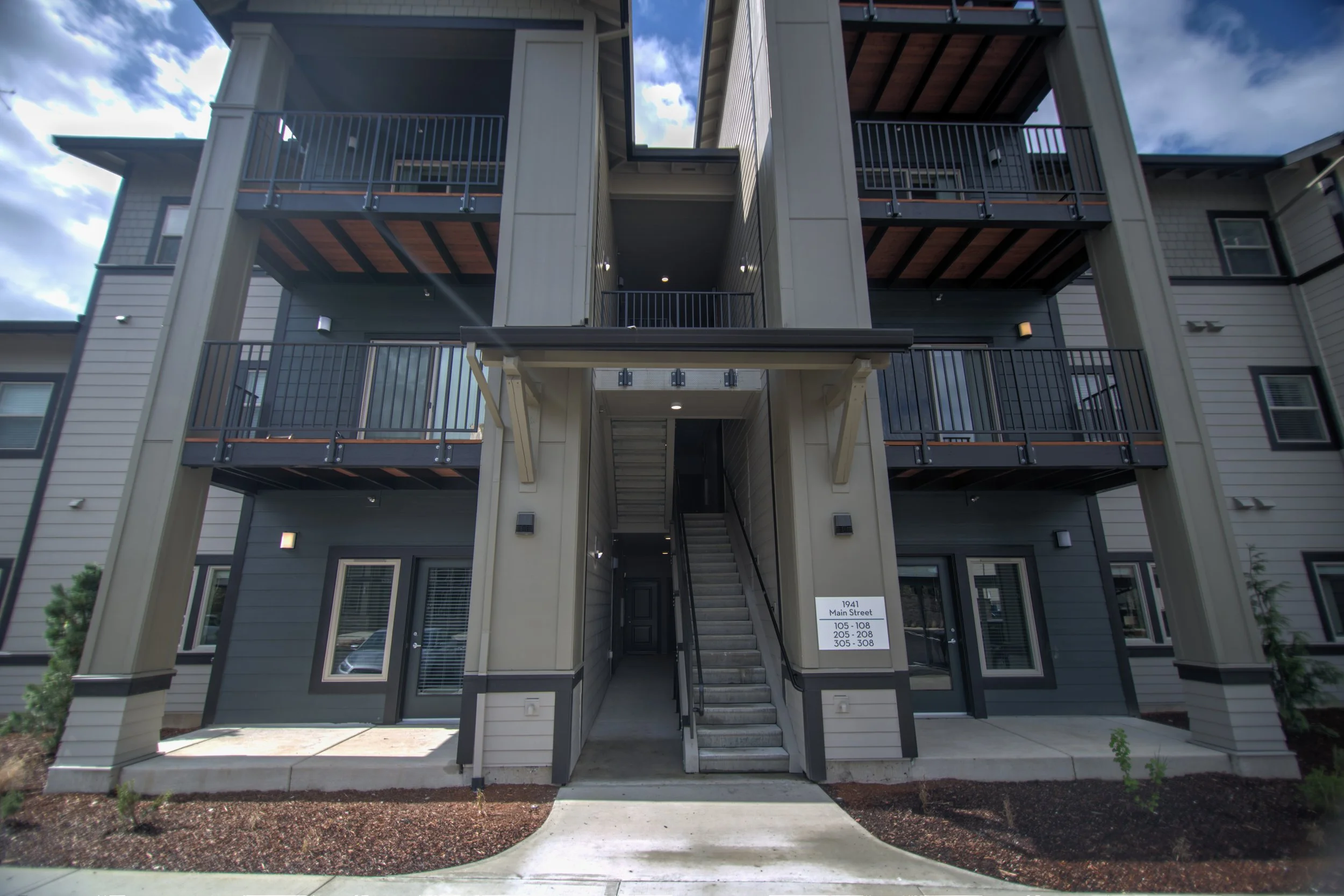 Exterior view of a modern multi-story apartment building with balconies, stairs, and parking area, featuring gray and beige siding, and a walkway leading to the entrance.