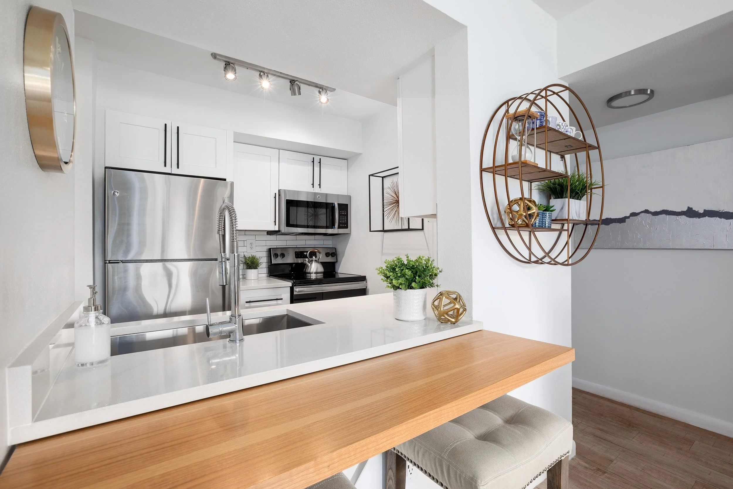 Modern kitchen with white cabinets, stainless steel refrigerator, microwave, black stove, and white countertop. Decor includes green plants, decorative objects, and a wall clock.