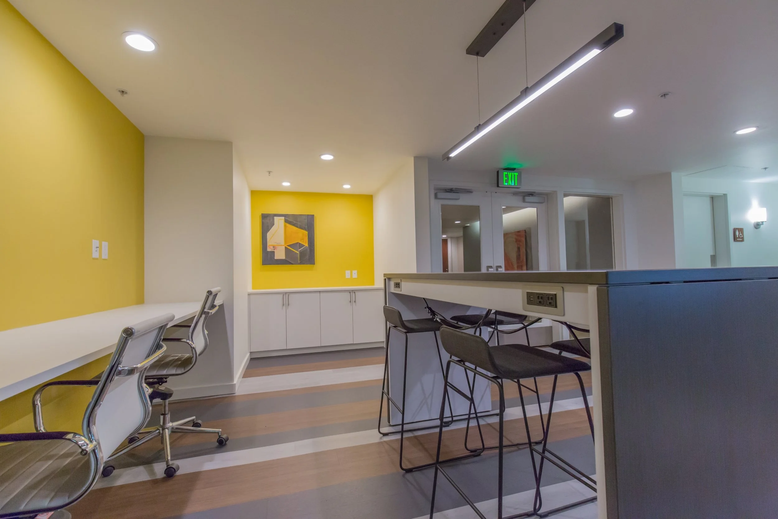 Empty modern office or coworking space with yellow accent walls, gray and white furniture, including chairs and a counter, with artwork on the walls, and ceiling lights.