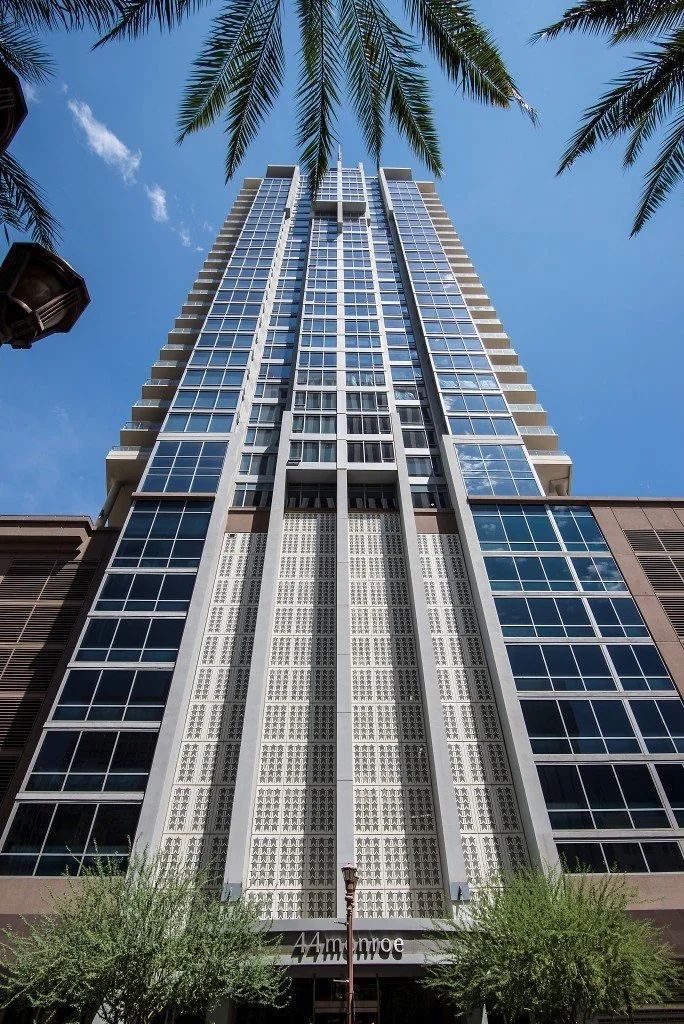Looking up at a tall modern high-rise building with glass windows, framed by palm trees and blue sky.