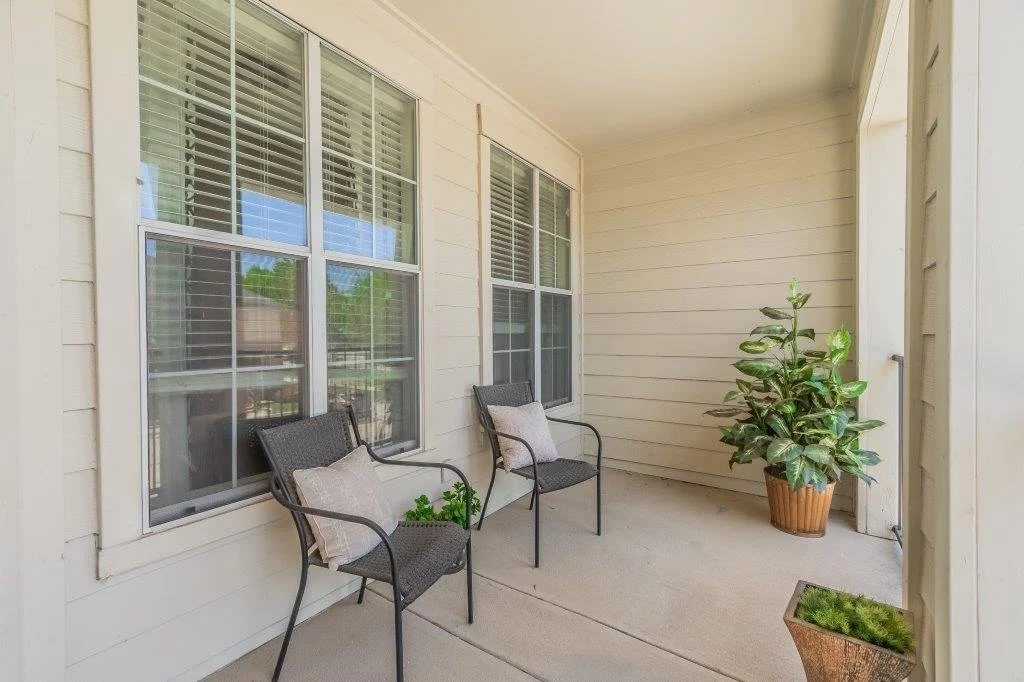 An outdoor patio with two black chairs, beige pillows, a potted plant on the floor, and a large green leafy plant in a pot, adjacent to beige siding and windows.