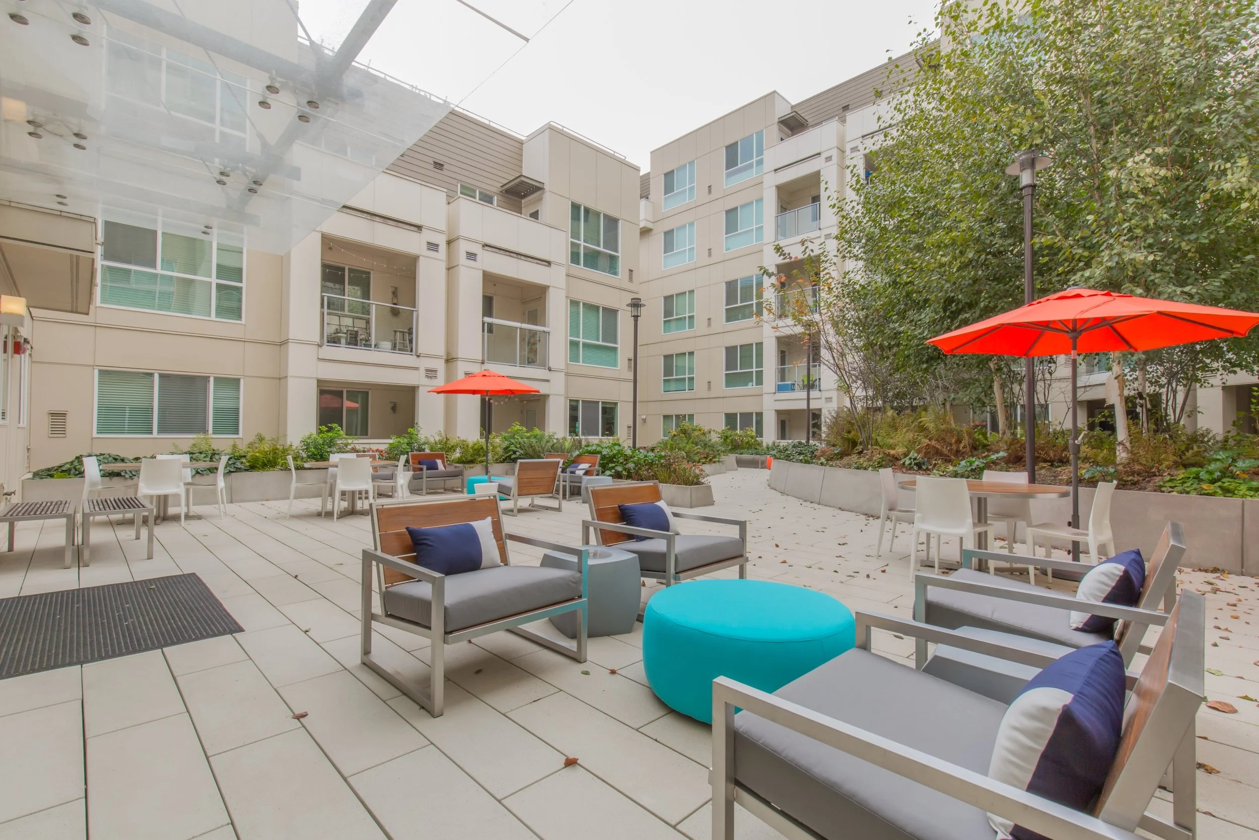 Outdoor patio area in a modern apartment complex with chairs, tables, colorful umbrellas, and trees.