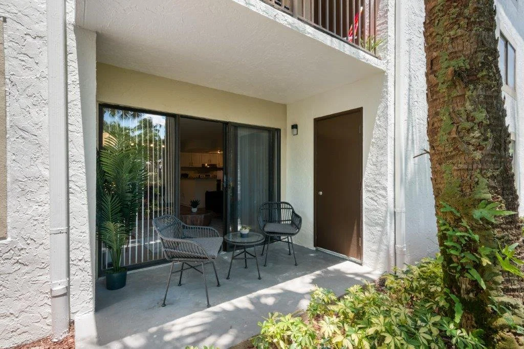 Outdoor patio with two black chairs, a small black table, a potted plant, adjacent to sliding glass door and a brown door, with a large tree and plants in the foreground.