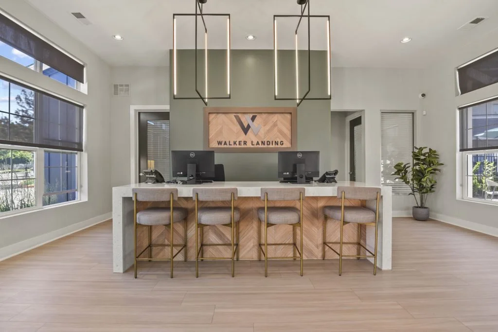 Modern reception area with a white marble counter, five cushioned bar stools, desktop computers, and a Walker Landing sign on the wall behind the counter. Large windows with blinds, corner plant, and minimalistic lighting.