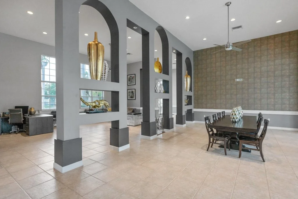 Interior of a modern dining area with a dark wooden table and six matching chairs. Decorative vases are placed on the table. The room features large windows, a textured accent wall, and a ceiling fan. Ornamental shelves with gold vases and sculptures