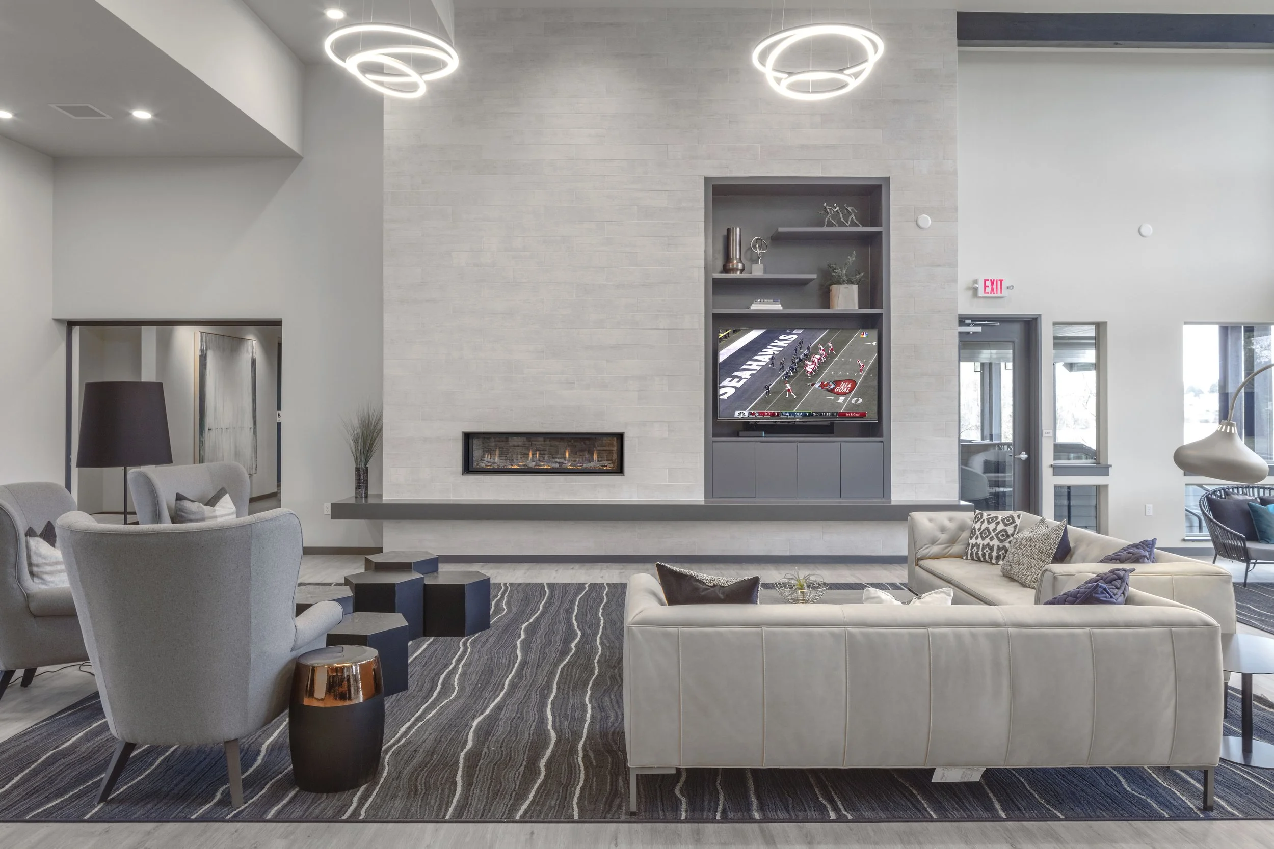 Modern living room with gray and white furniture, a fireplace, a wall-mounted TV showing a football game, and contemporary lighting fixtures.