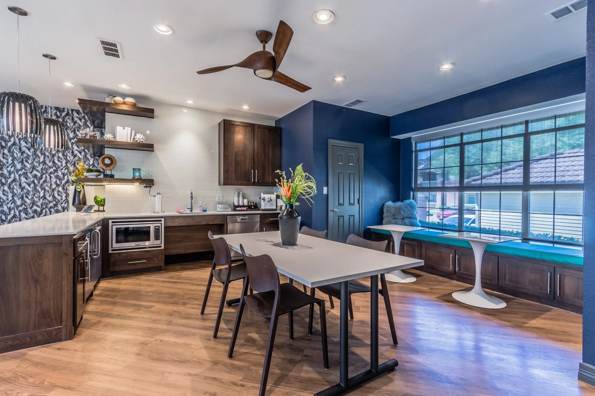 Modern kitchen and dining area with wooden flooring, dark wood cabinets, a white table with chairs, a large window with a built-in window seat, and decorative shelving on the wall.