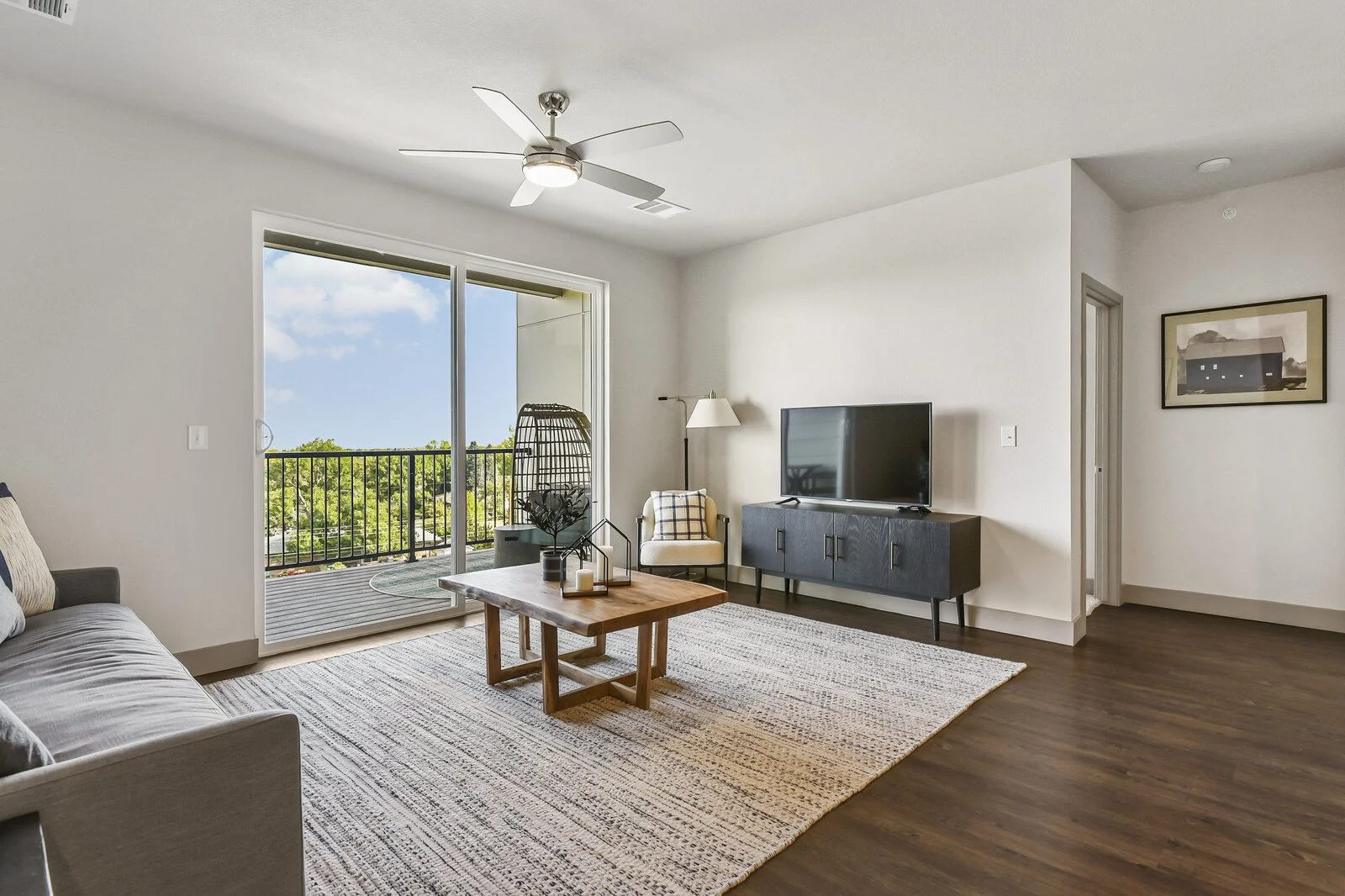 Modern living room with sliding glass door leading to balcony, ceiling fan, flat-screen TV on black cabinet, armchair, sofa, coffee table, and framed picture on wall.