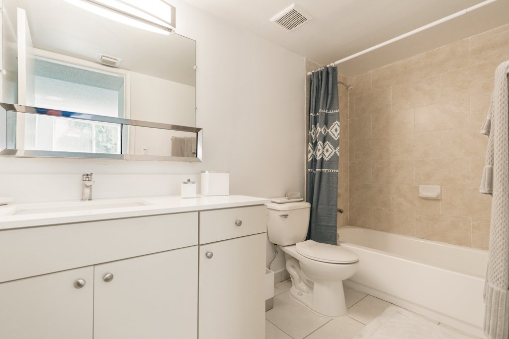 A clean bathroom with a white vanity, large mirror, toilet, bathtub with beige tiles, and a gray shower curtain.