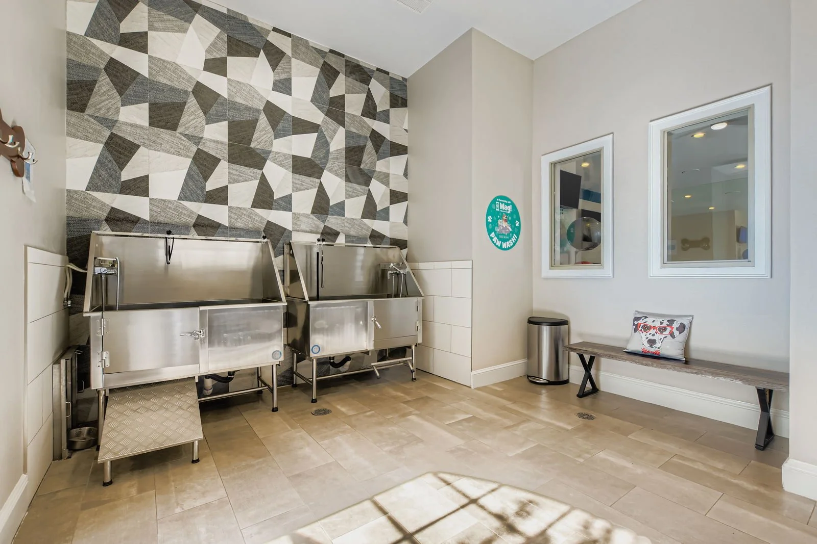 Dog wash station with two stainless steel tubs, a bench with a pillow, and a decorative tiled wall in the background.