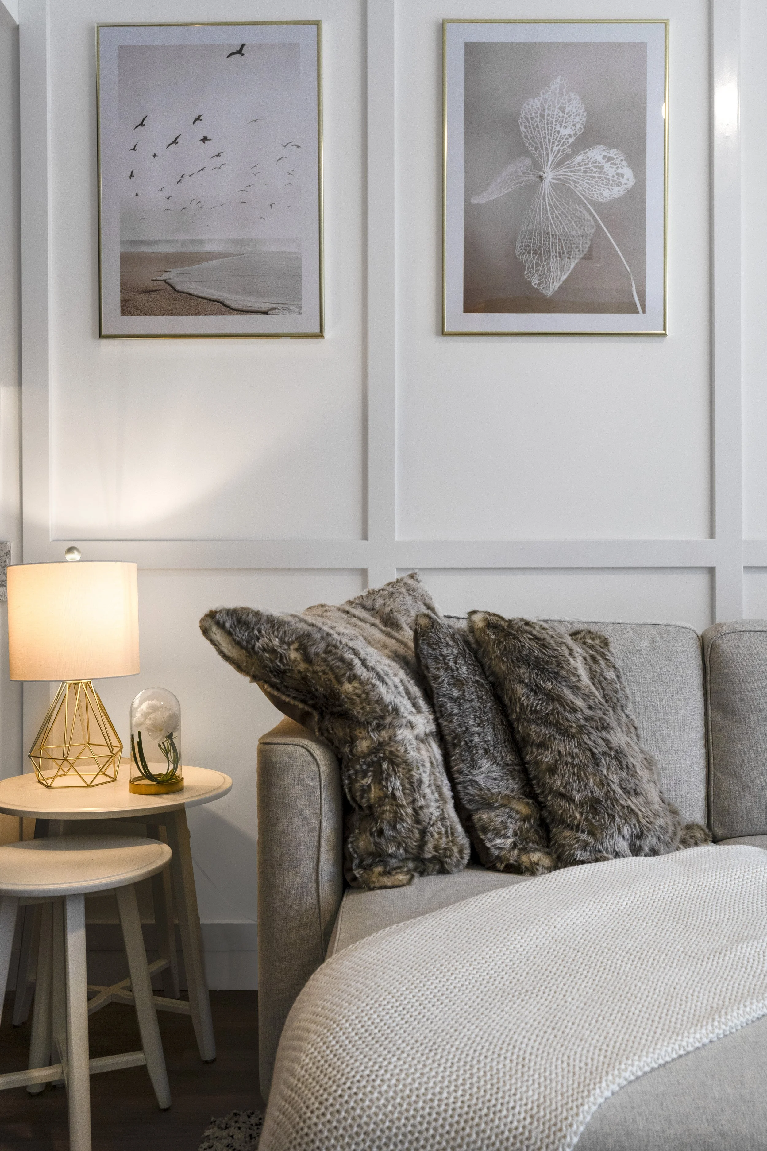 A cozy living room corner with three faux fur pillows on a beige sofa, a side table with a lamp and decorative terrarium, and two framed art prints of a beach scene and leaf skeleton on the white paneled wall.