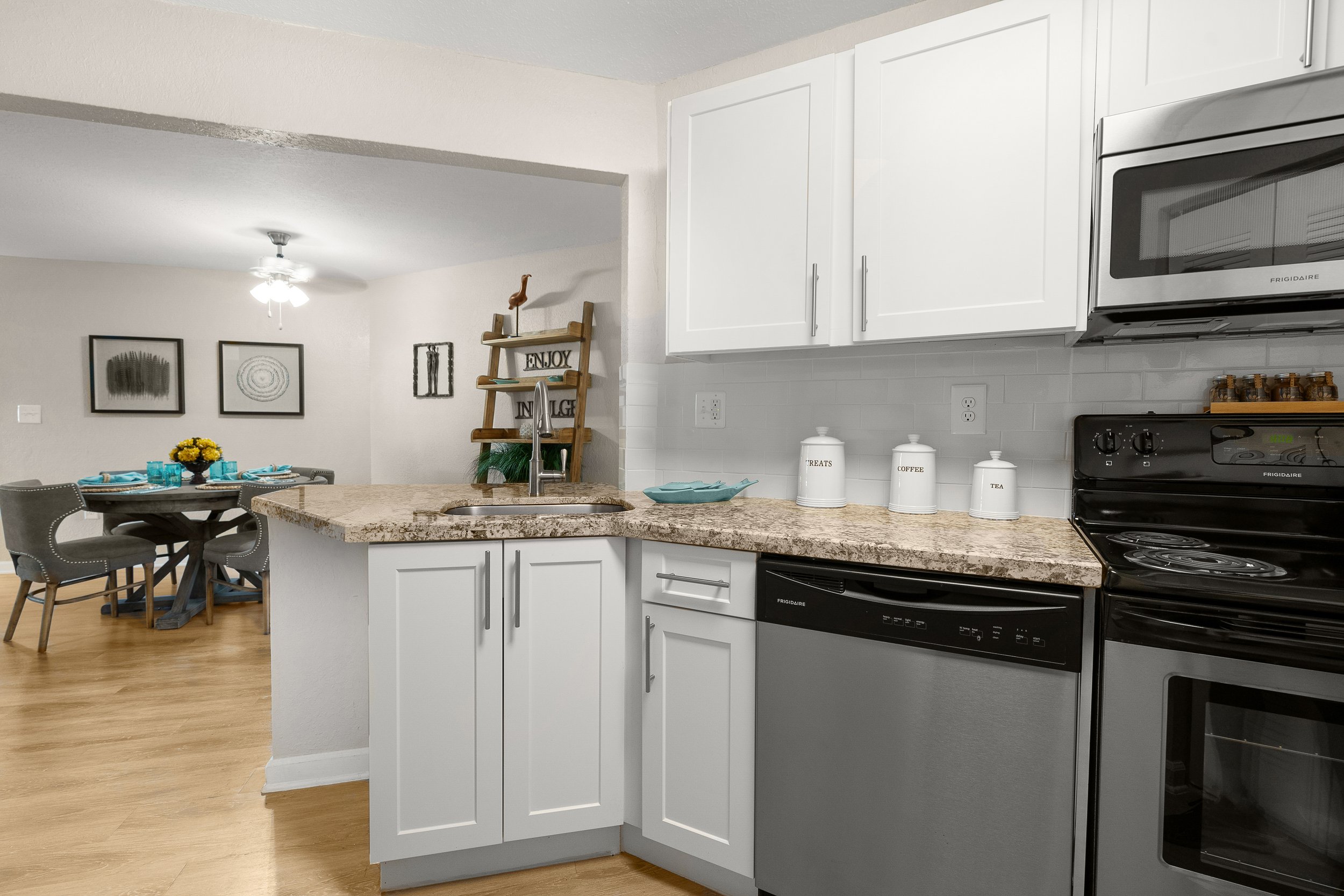 View of a modern kitchen with white cabinets, granite countertops, stainless steel appliances, and a dining area with a round table set for four, decorated with a yellow floral centerpiece. Talavera jars labeled 'Treats,' 'Coffee,' and 'Tea' are on t