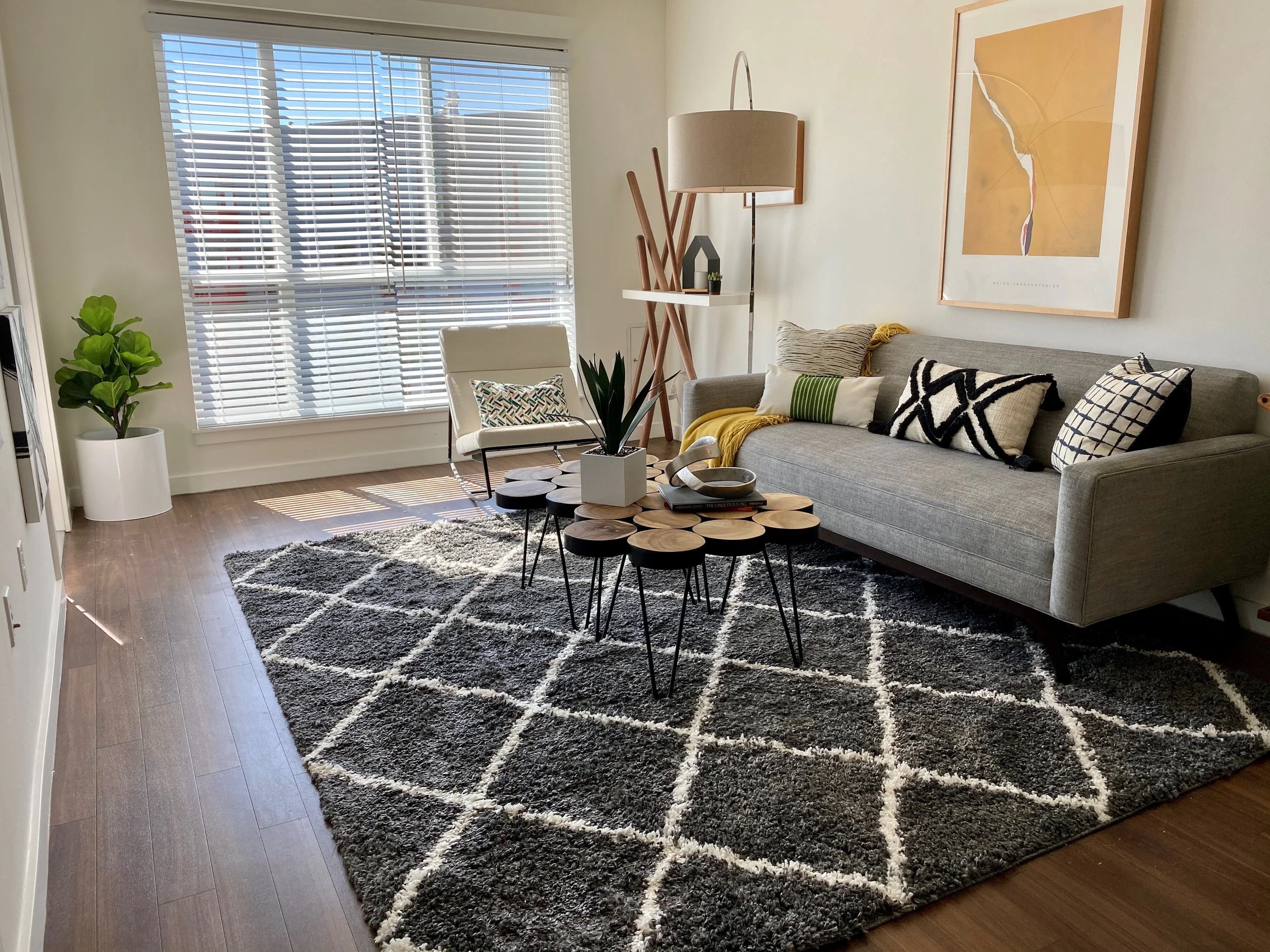 Living room with gray sofa, black and white cushions, a white chair, a large window with blinds, a potted plant, a patterned rug, a multi-level coffee table, a wooden floor, a tall floor lamp, and framed artwork on the wall.