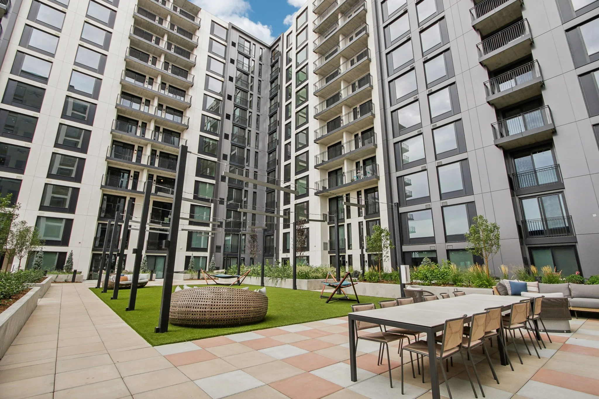 View of a modern apartment courtyard with outdoor furniture, a dining table with chairs, lounge seating, green landscaping, and tall apartment buildings surrounding the area.