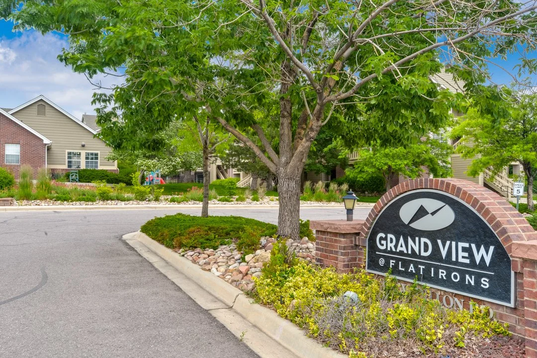 Entrance sign for Grand View at Flatirons residential community, with a brick arch, a small lamp post, and trees with green leaves surrounding the area.